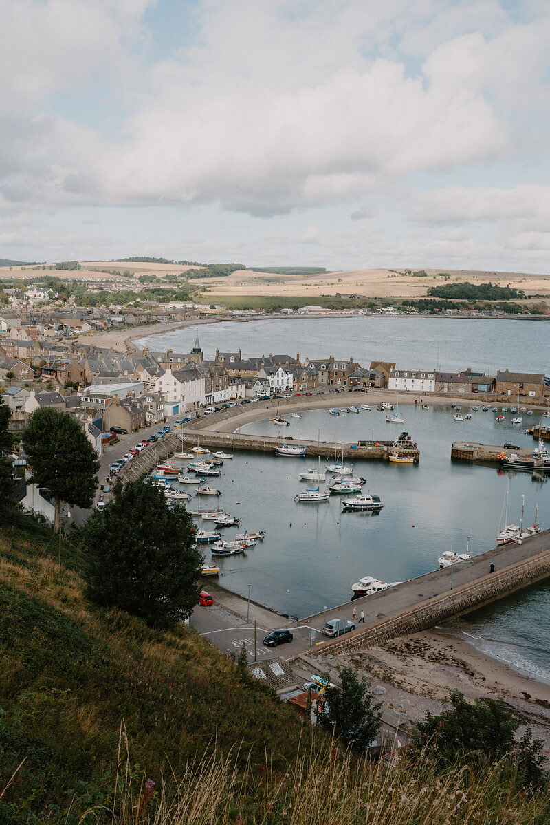 Boats crowd a stone harbour in a small coastal town, pastel houses lining the water and a sandy beach beyond.