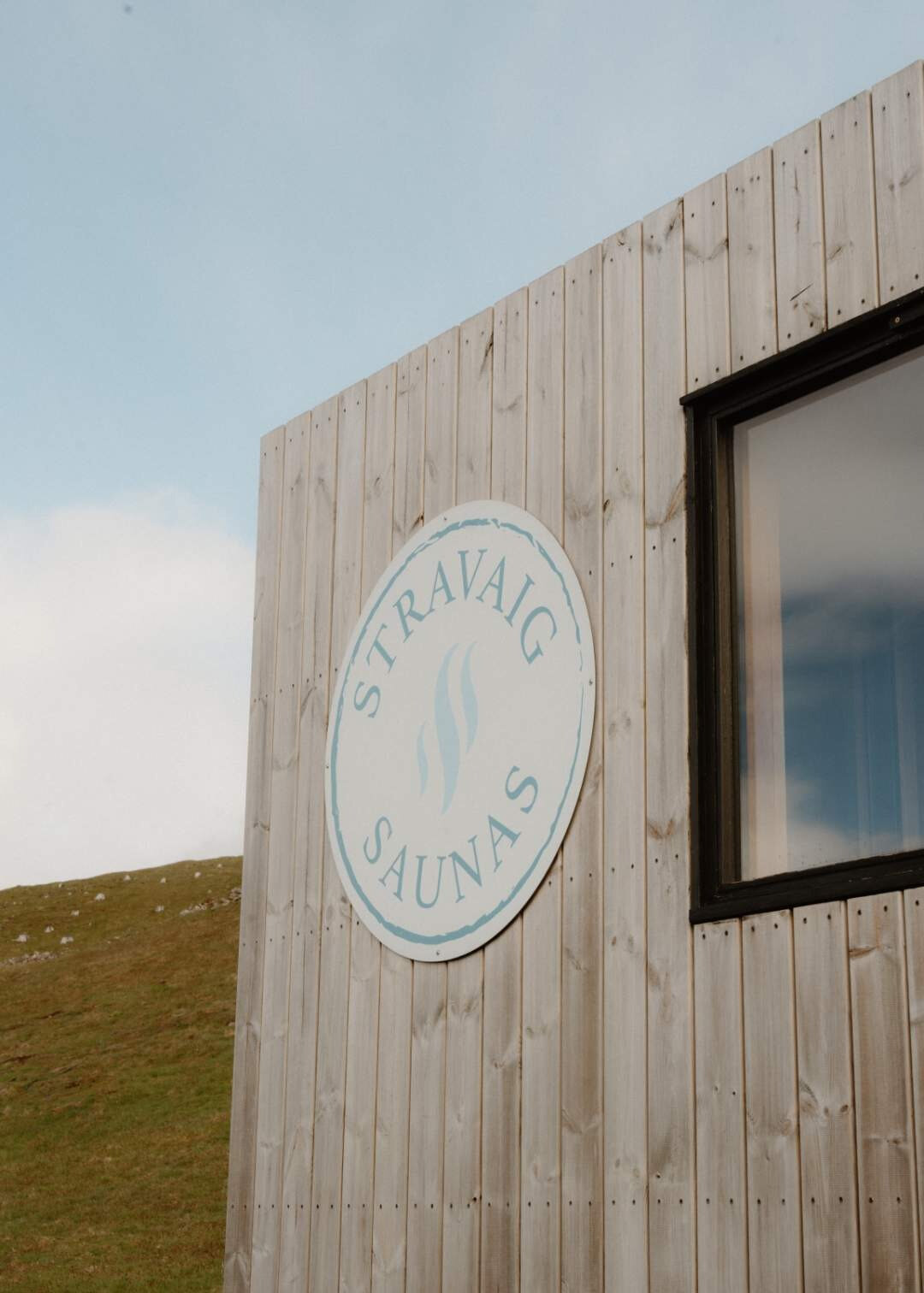 A circular Stravaig Saunas sign mounted on the timber cladding beside a window