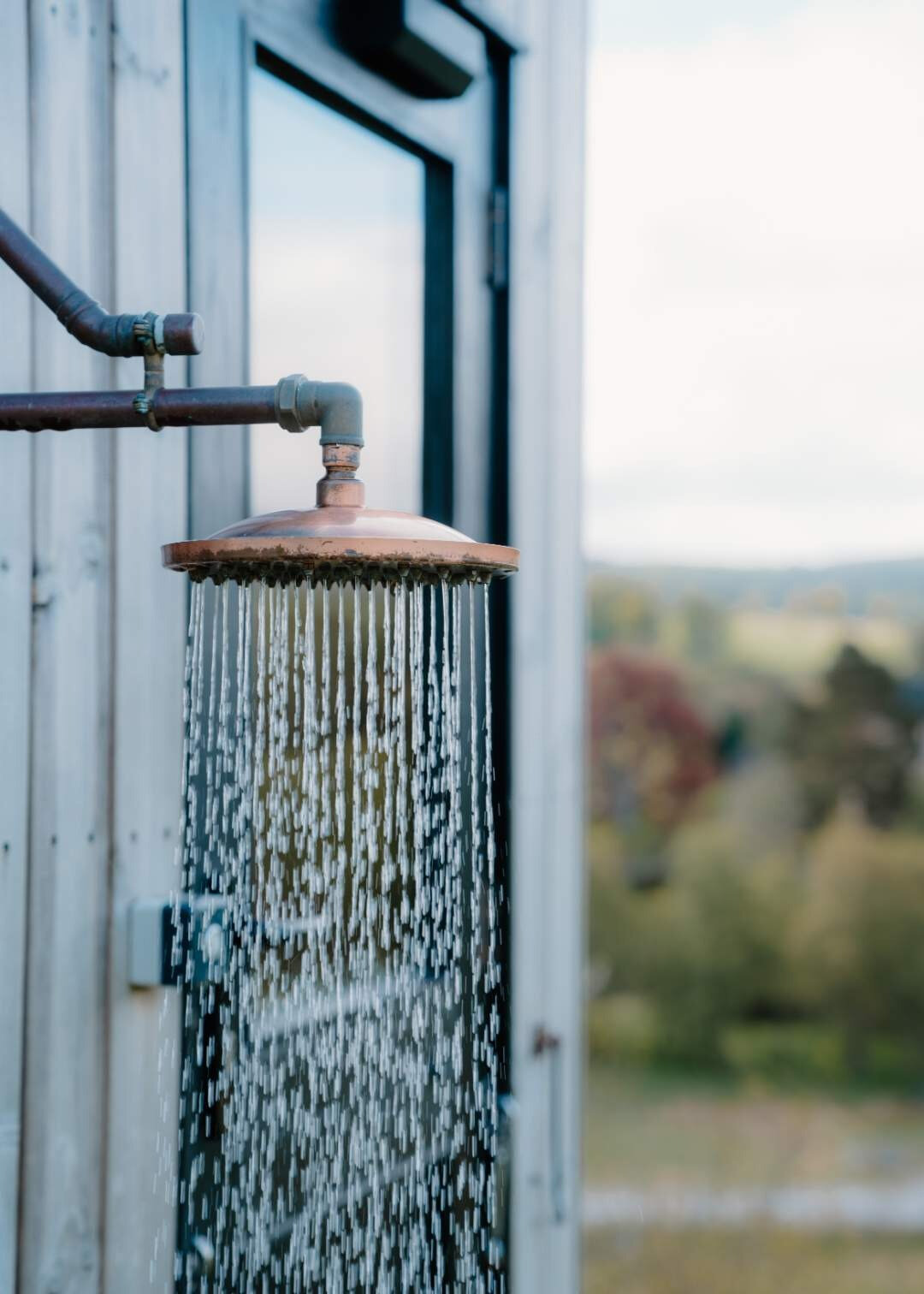 An outdoor copper showerhead runs beside a timber cabin, hills stretching into the distance, at Stravaig Saunas
