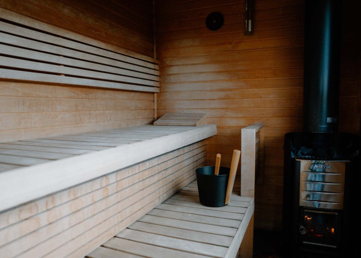 Slatted wooden benches line a sauna beside a black stove, a bucket and ladle set nearby, at Stravaig Saunas