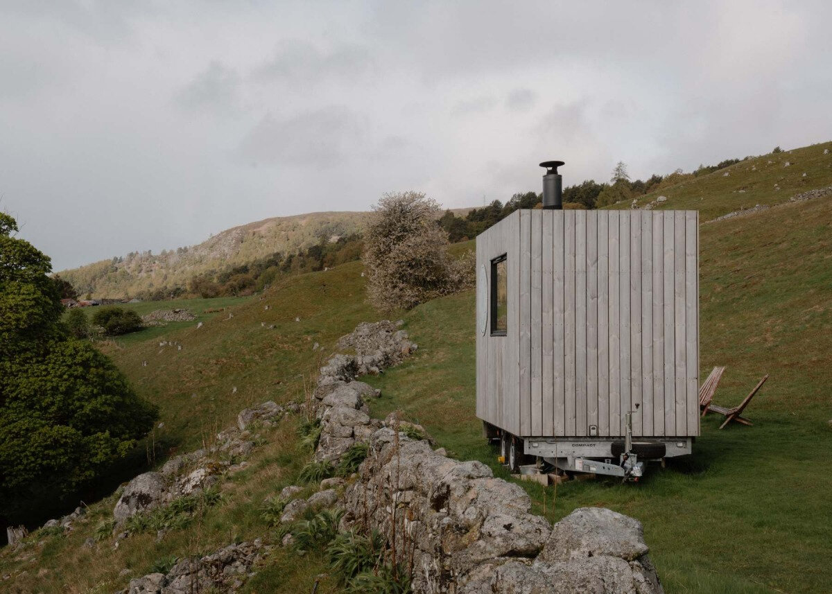 A timber sauna cabin with a stovepipe chimney stands on a grassy hillside beside a drystone wall, deck chairs alongside, at Stravaig Saunas