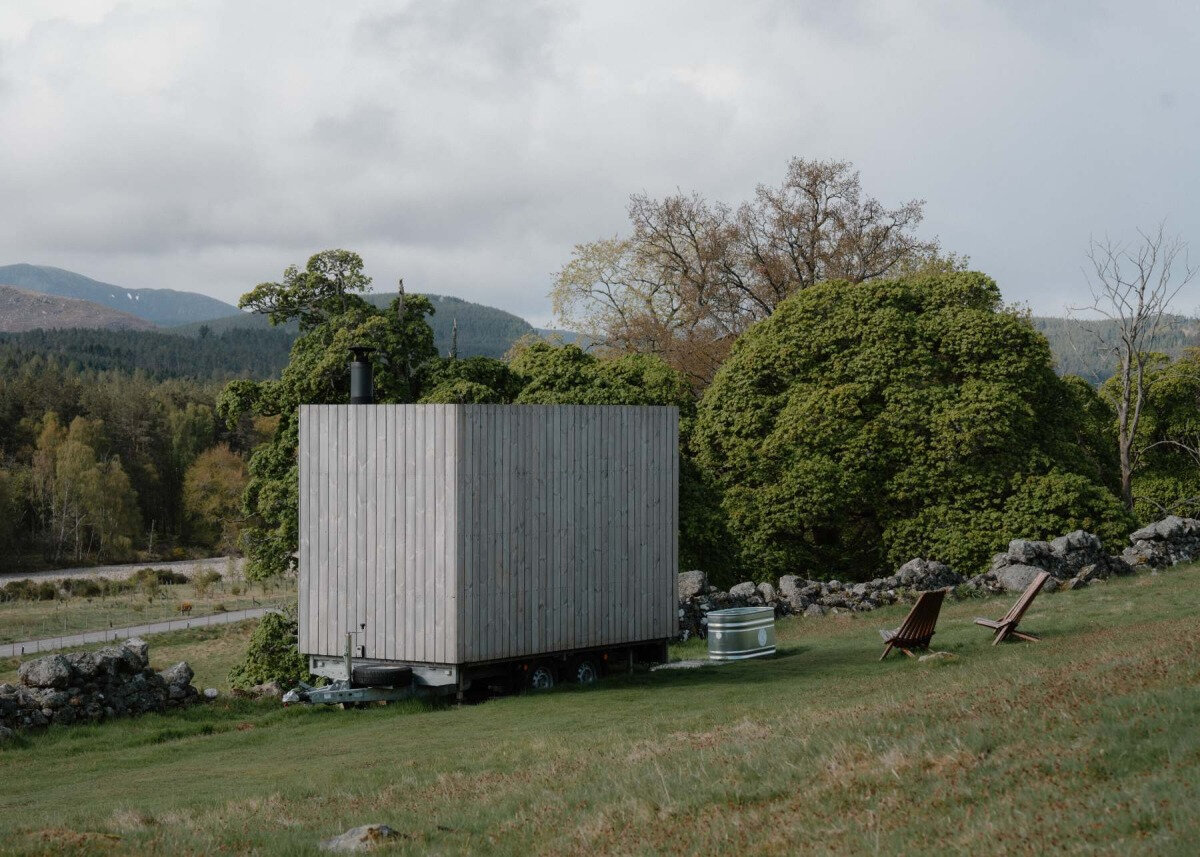 A timber-clad mobile sauna parked on grass, with a cold-plunge tub and deckchairs beside it