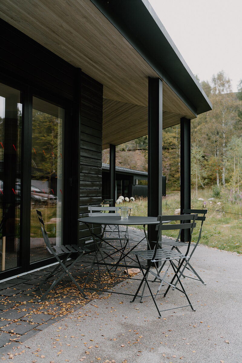 A modern black-clad building's covered patio holds a bistro table and chairs, forest rising behind.