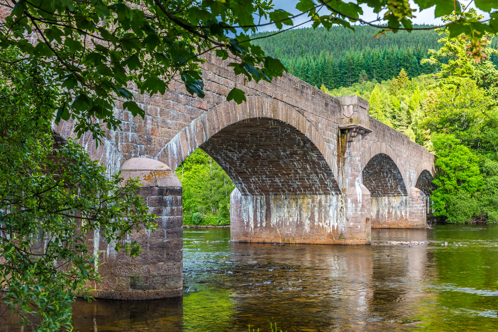 The granite bridge over the River Dee at Ballater, its arches reflected in the brown water, wooded hills behind and a branch hanging over the bank.