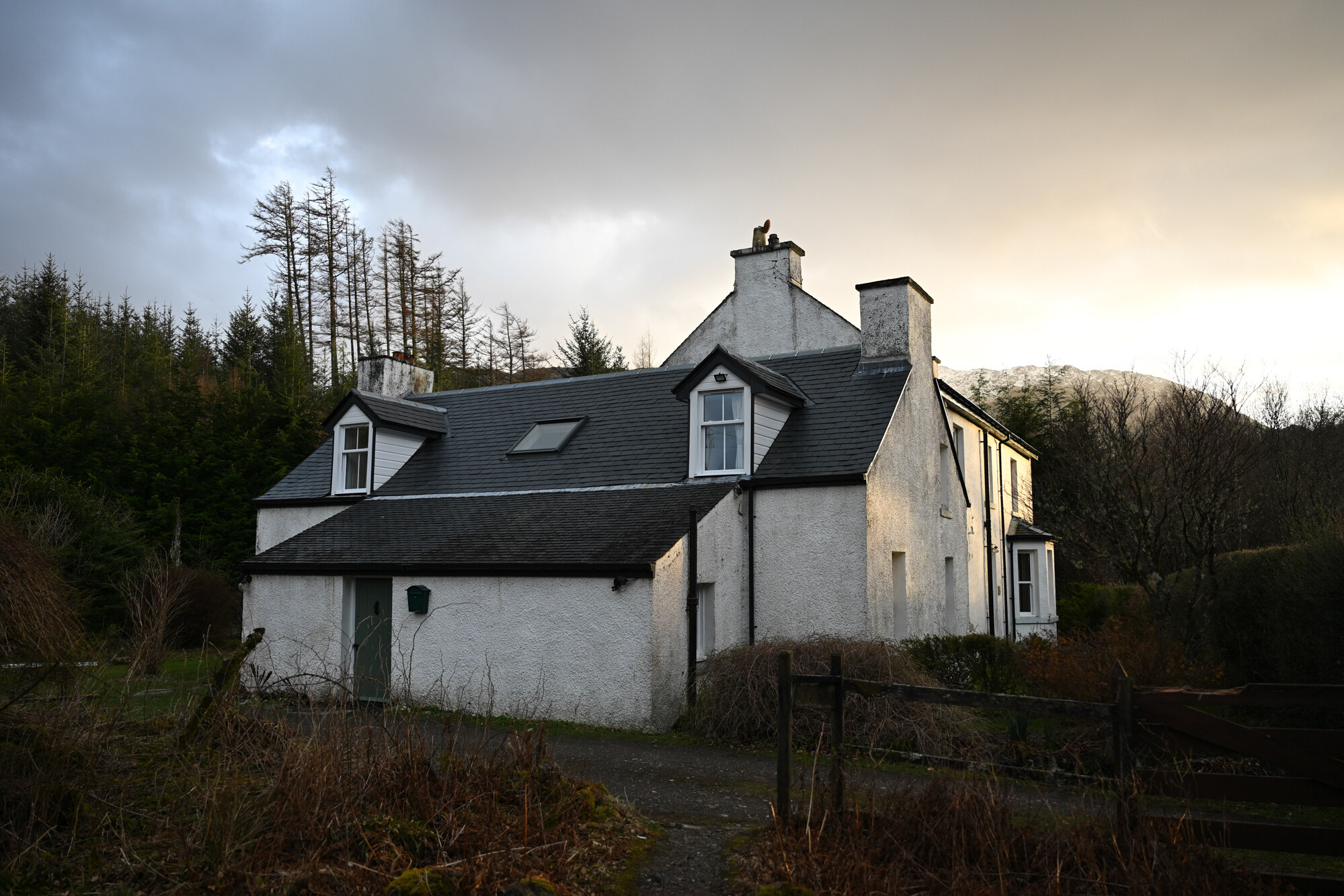 The white-harled lodge and its dormers under a pale sky, larches on the slope behind.