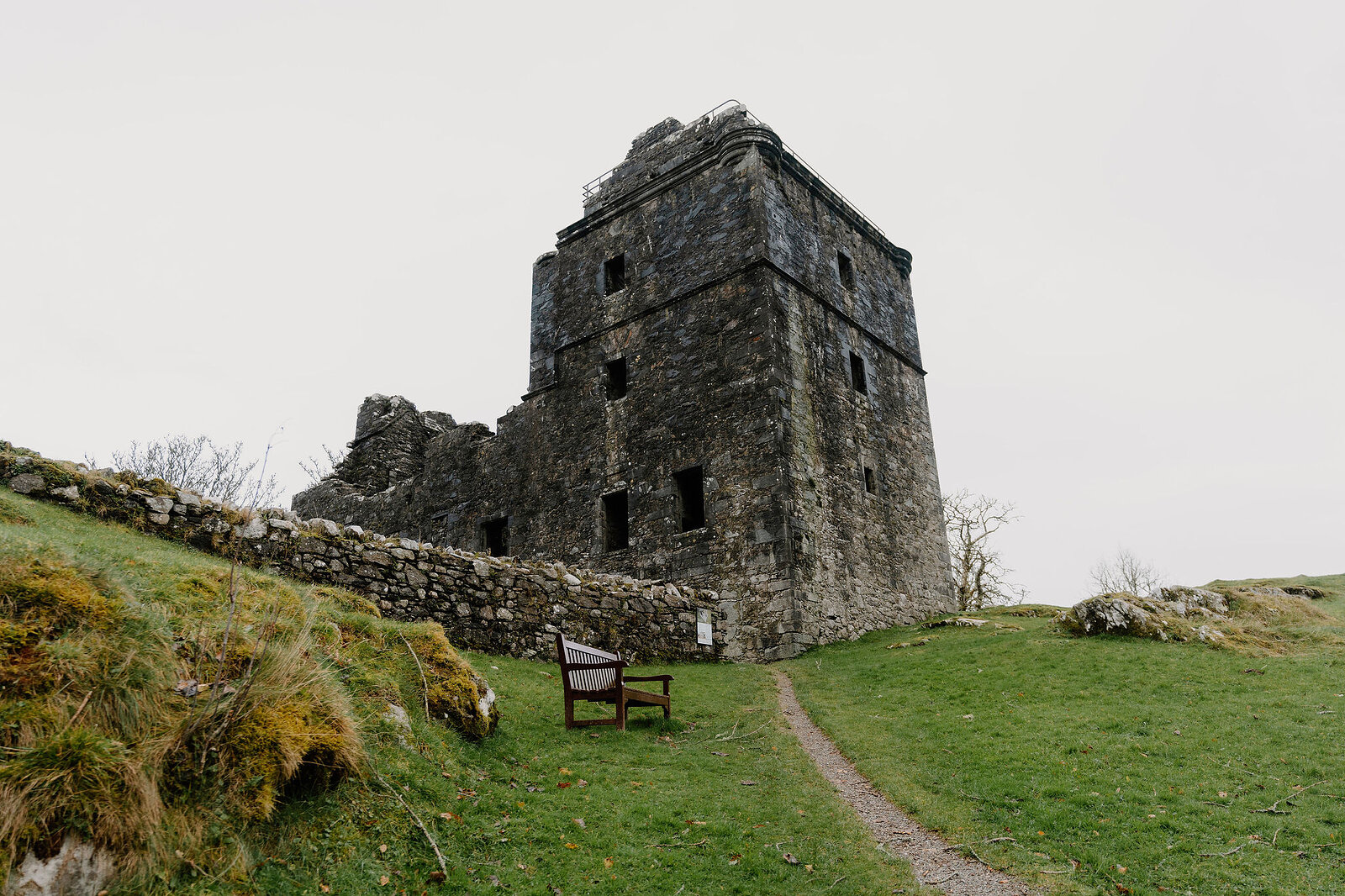 A ruined stone tower keep rising above a mossy dry-stone wall, a wooden bench set on the grass before it