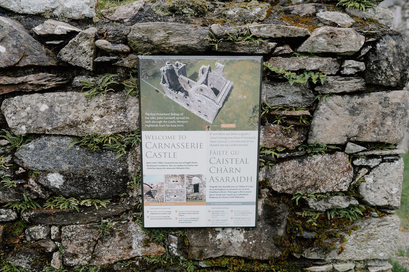 An information board reading Welcome to Carnasserie Castle mounted on a mossy stone wall