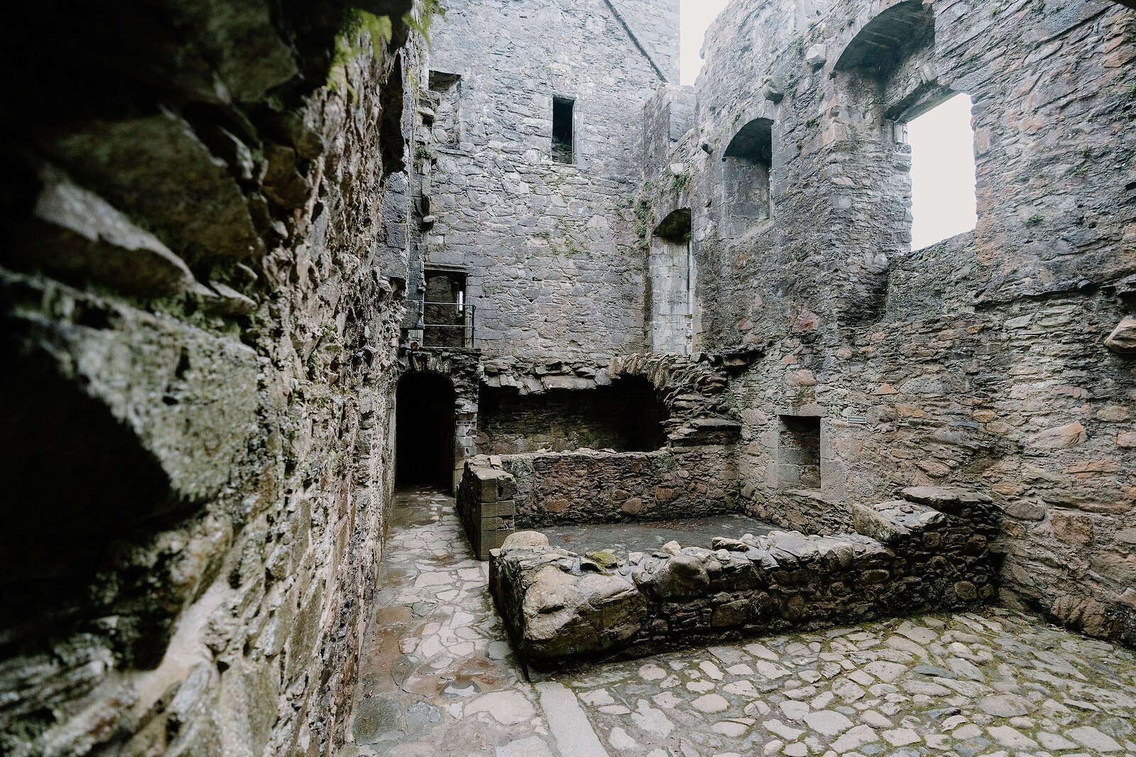 The roofless stone interior of a ruined castle, arched window openings and a raised hearth visible