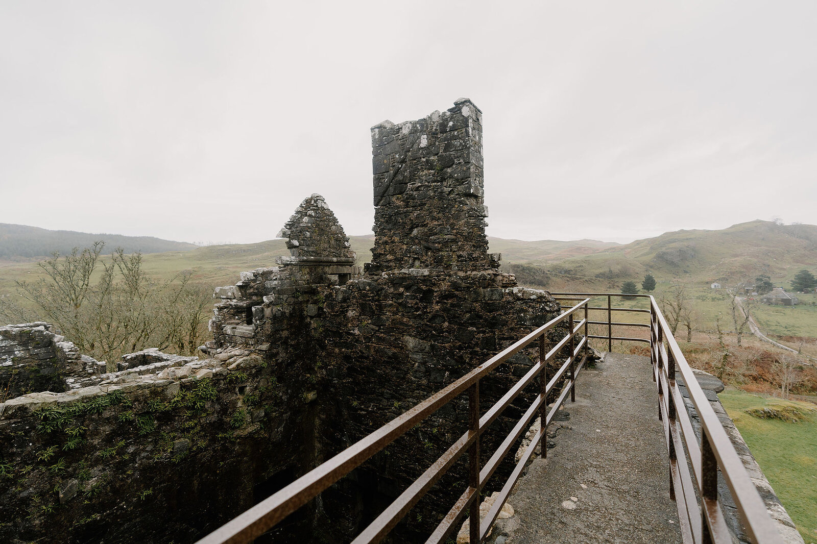 A ruined stone tower connected by a metal walkway, hills fading into the distance beyond