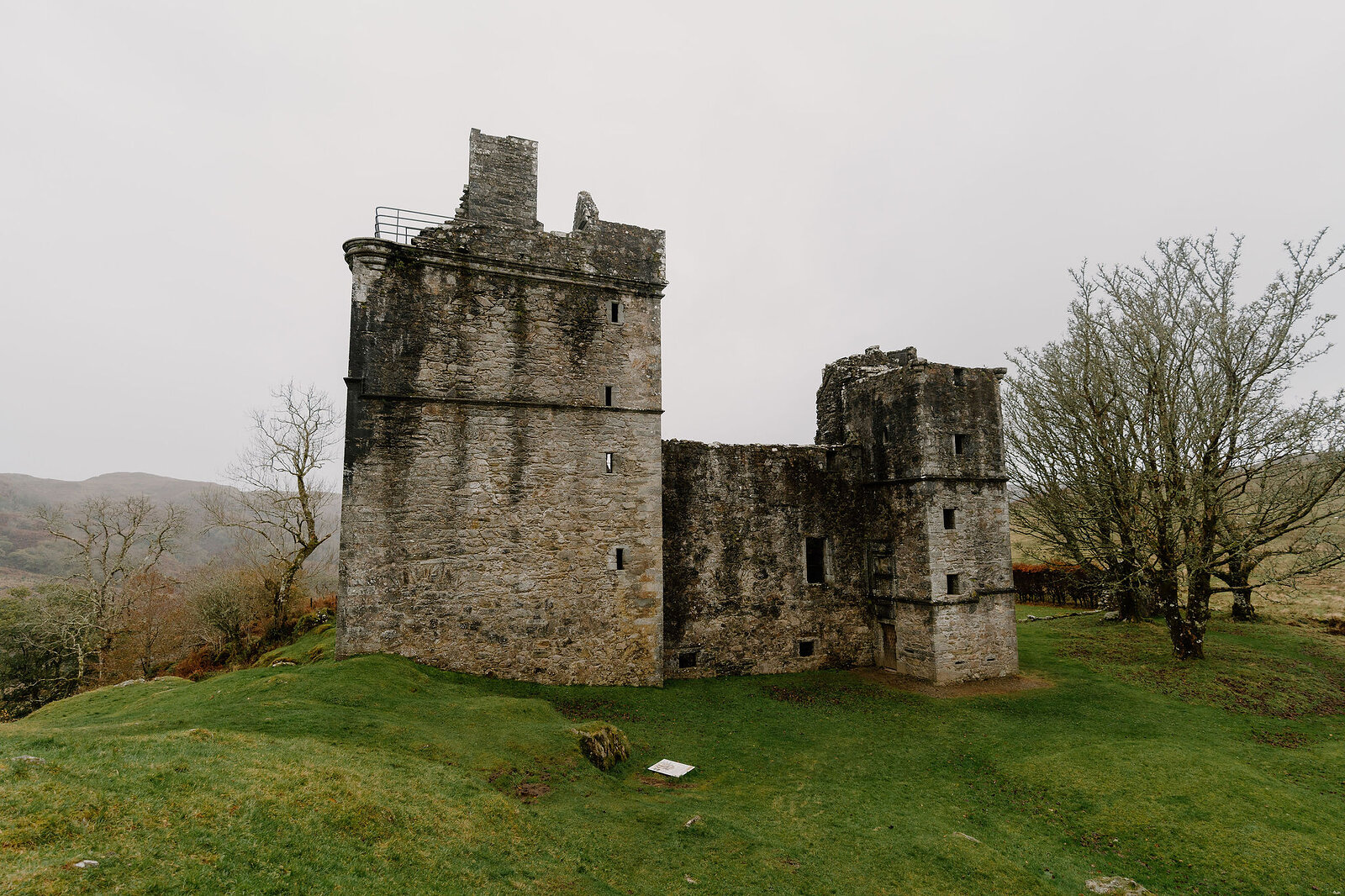 A ruined stone tower keep with a collapsed corner, trees framing it on a grassy mound