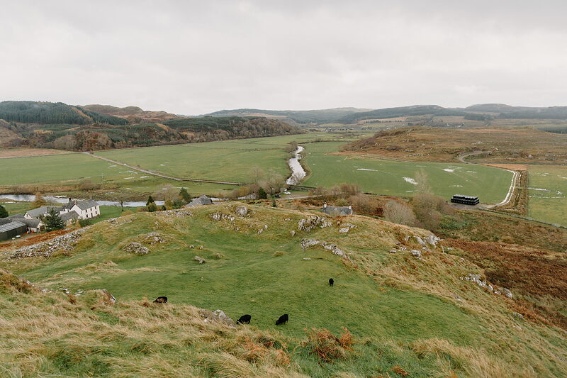 A ring of stones set on a grassy hillfort overlooking farmland and a winding river, cattle grazing below.