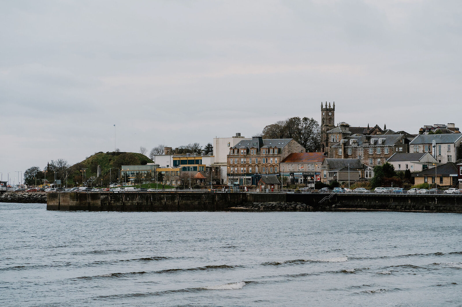 A grassy mound and church spire rising above a row of coastal buildings, waves lapping the shore below