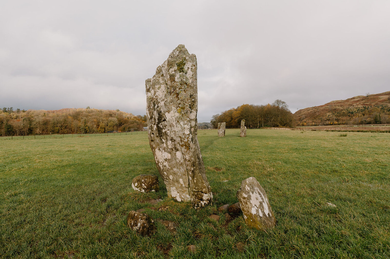 A tall standing stone flanked by two smaller stones in a grass field, hills in the distance