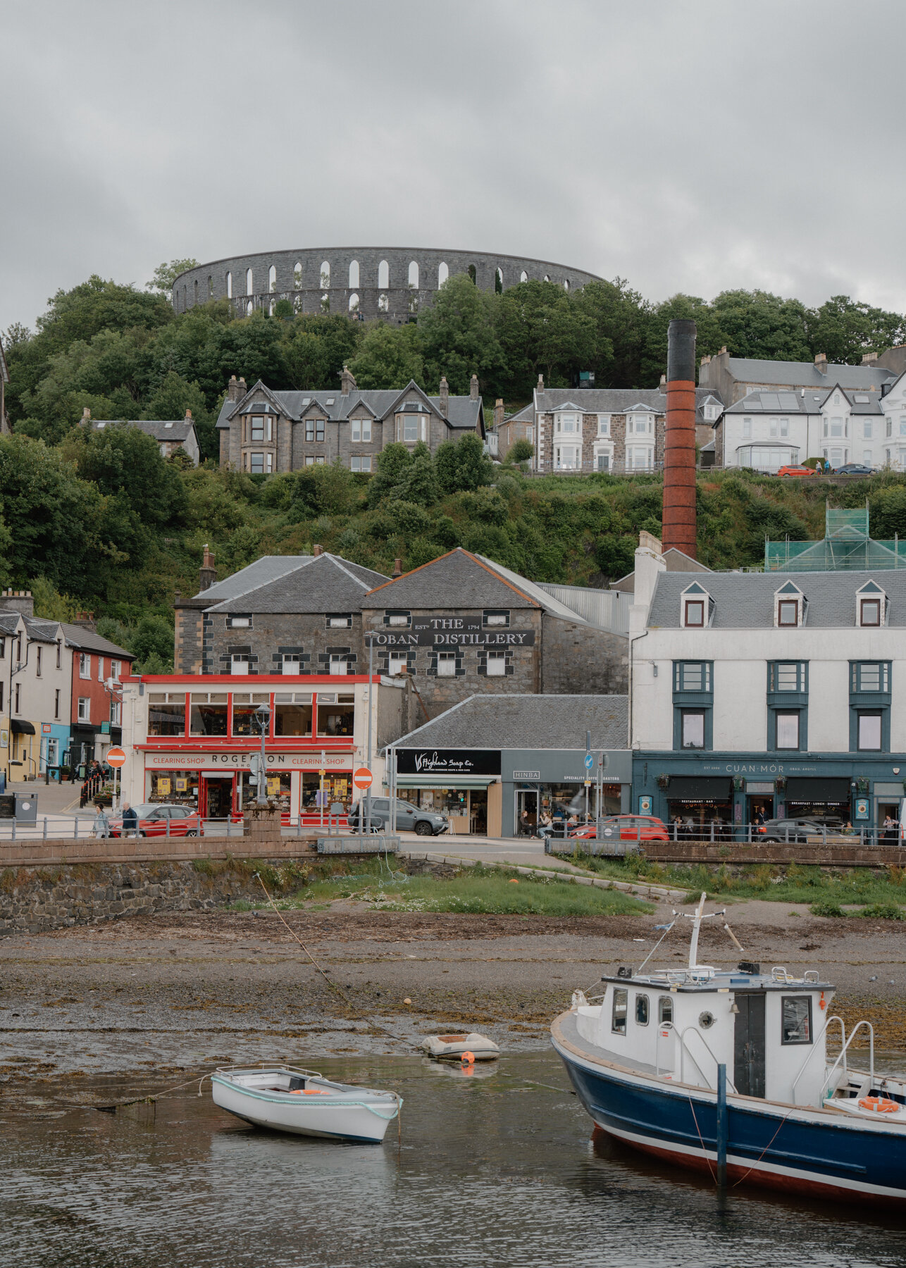 A hillside town rising below a ruined circular monument, shopfronts and fishing boats lining the harbour front.