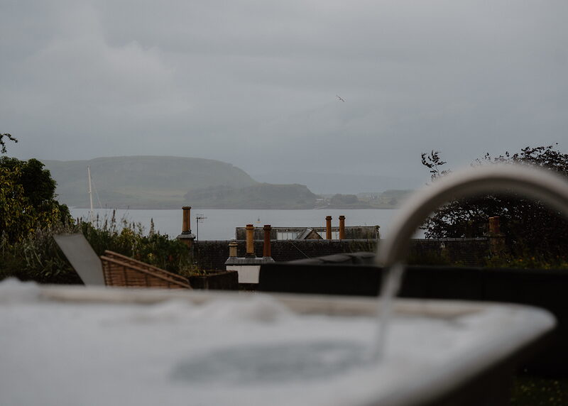 An outdoor stone bathtub filling with water on a terrace, a rocky coastal headland visible across the water.
