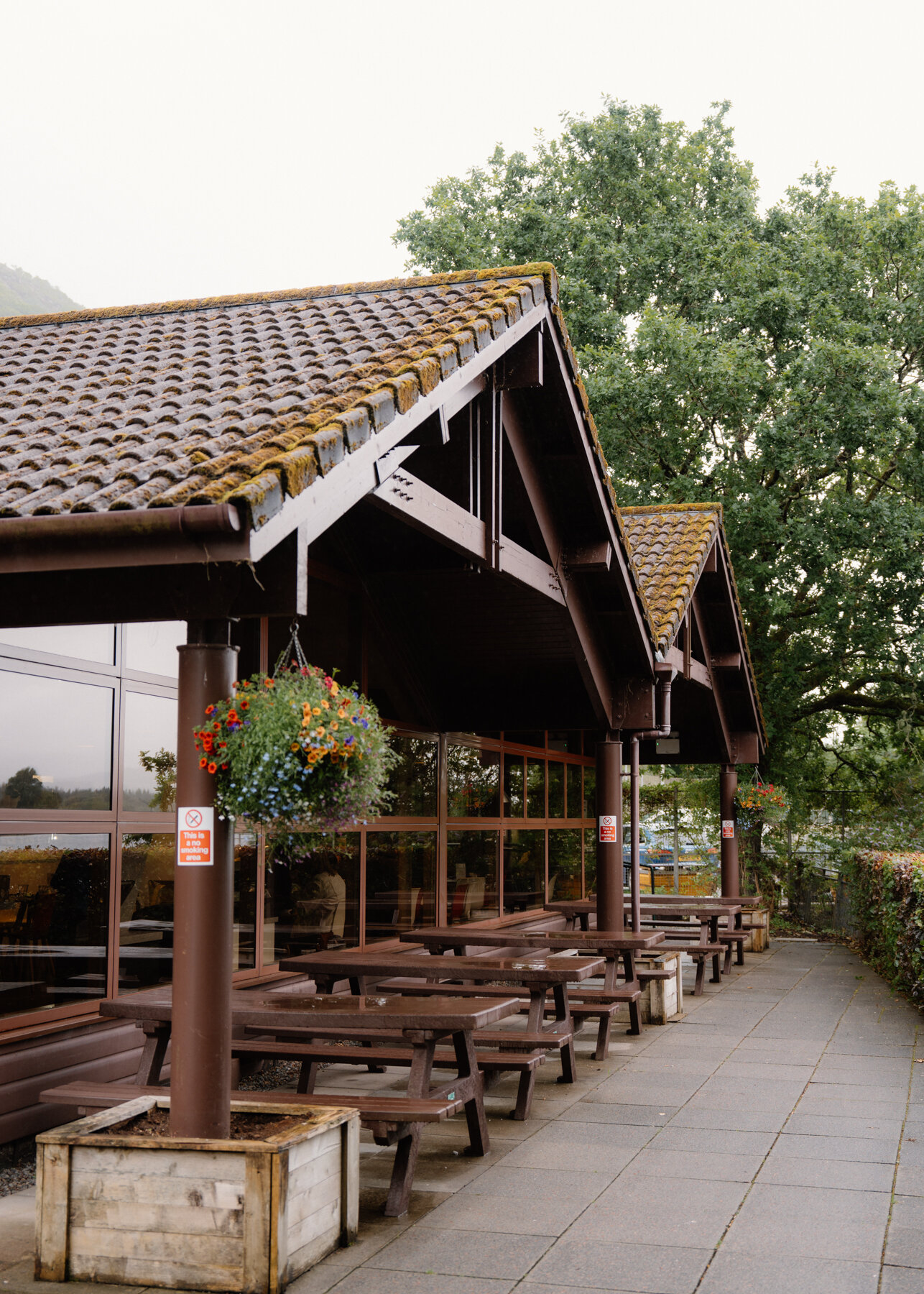 Hanging flower baskets hang from a covered timber walkway lined with picnic tables.