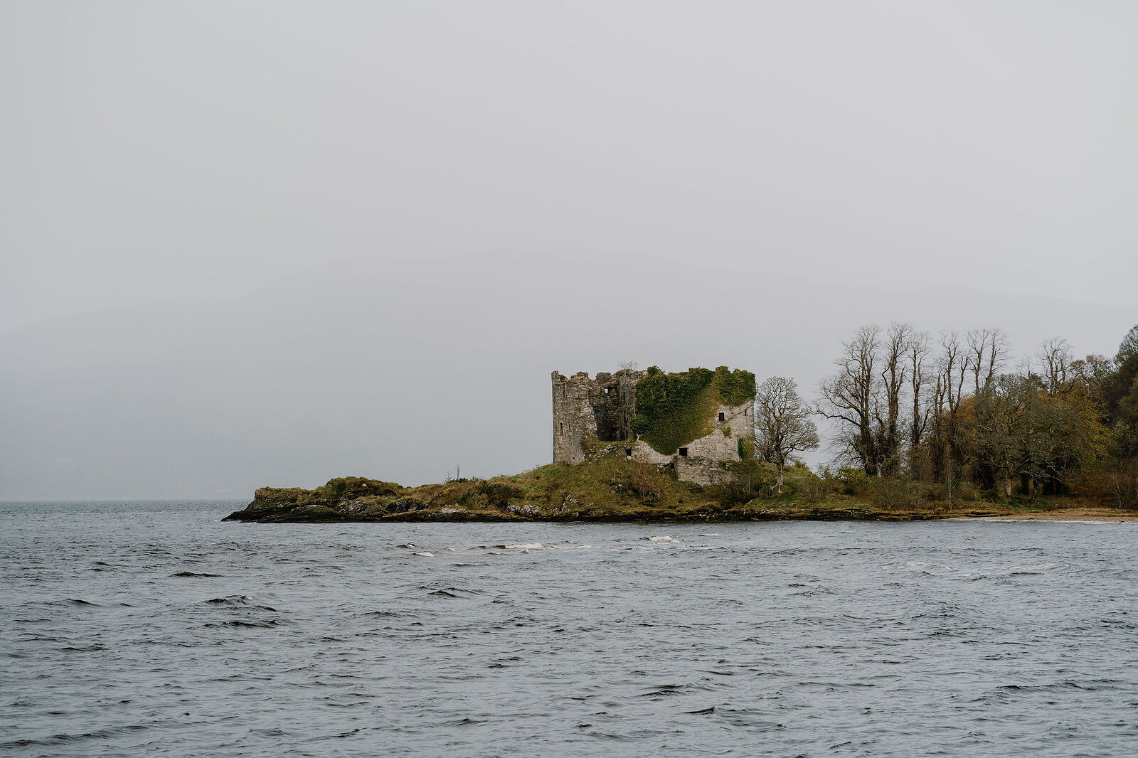 A ruined castle standing on a small tree-covered island in a loch, waves lapping the shore