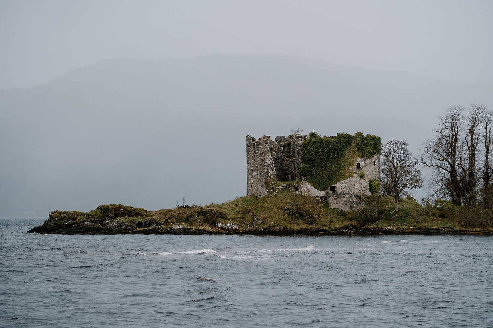The ruined castle on its low point of rock, ivy over one wall, grey water in front and hills in the mist.
