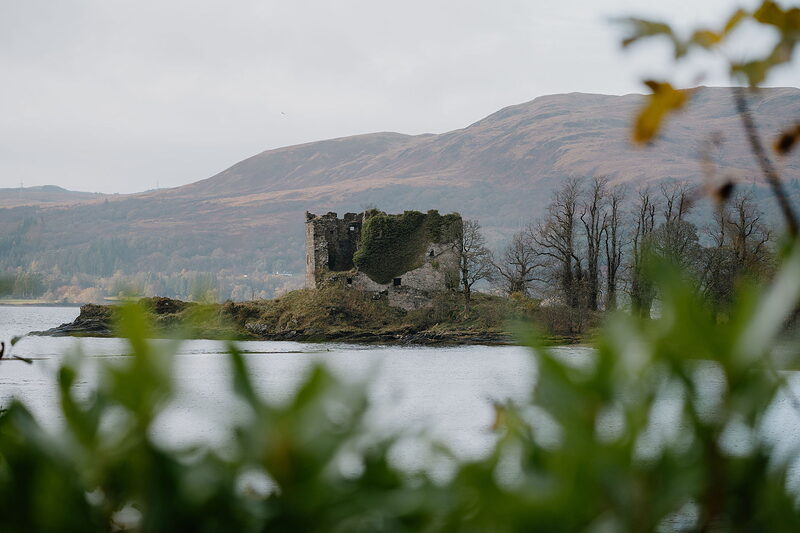 A ruined castle on an island across the loch, framed by foliage with hills behind.