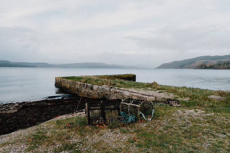A stone pier with creels stacked at its base, reaching into a calm loch bordered by hills.