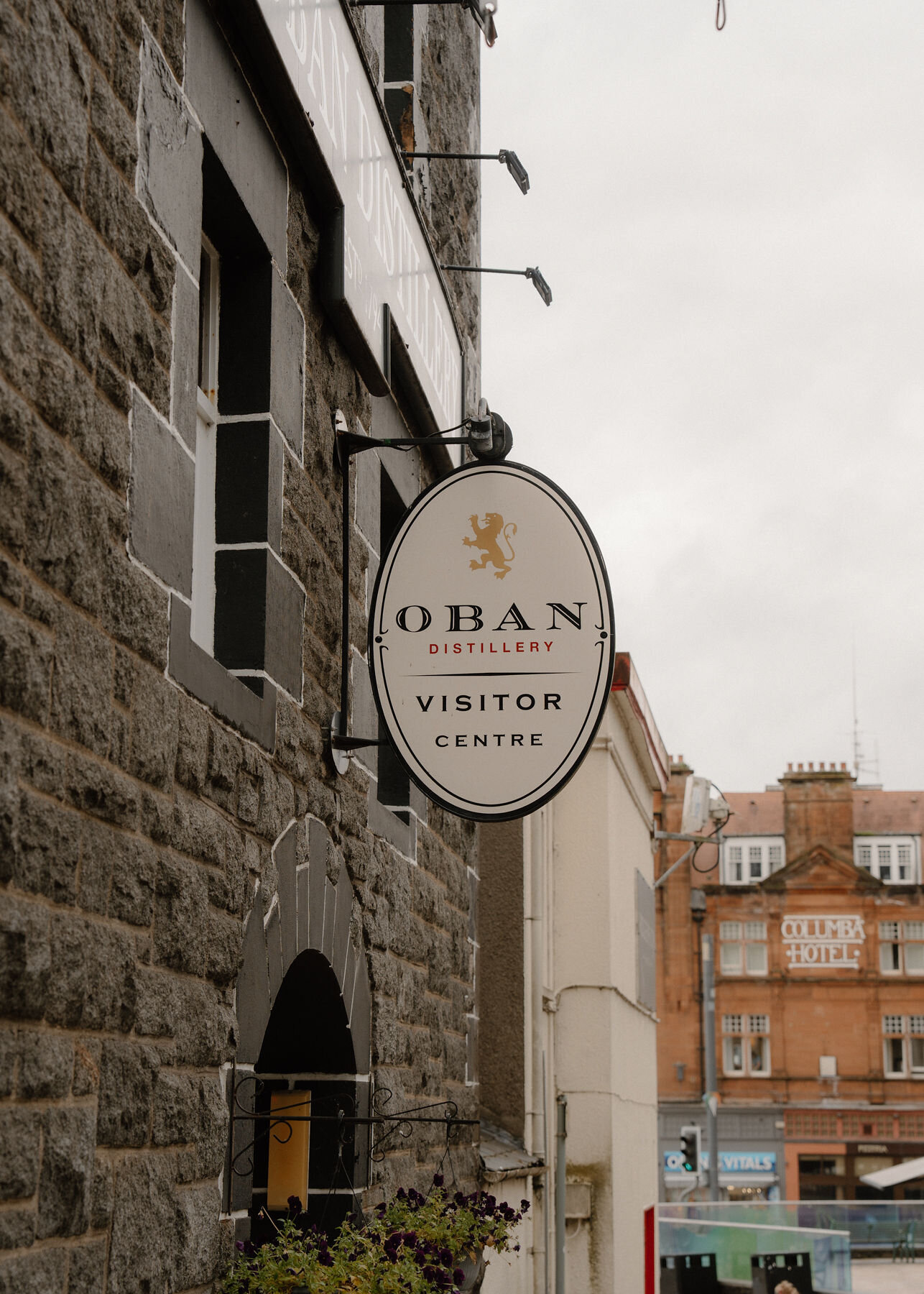 The oval Oban Distillery Visitor Centre sign hanging from the dark stone wall of the distillery.