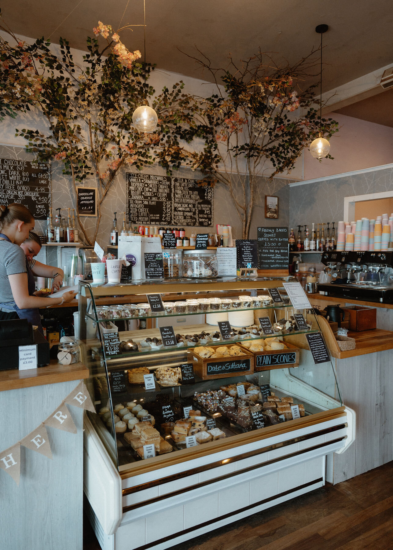 Oban Chocolate Company's counter under blossoming branches, cabinets of cakes and scones, a woman serving.