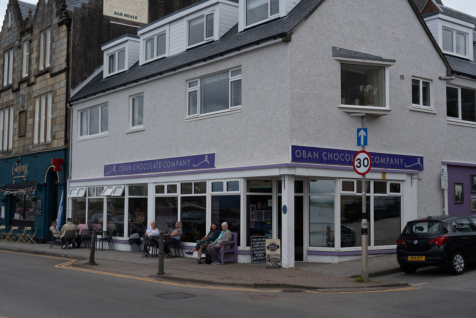 Oban Chocolate Company on its corner of the Corran Esplanade, people on benches outside its long windows.