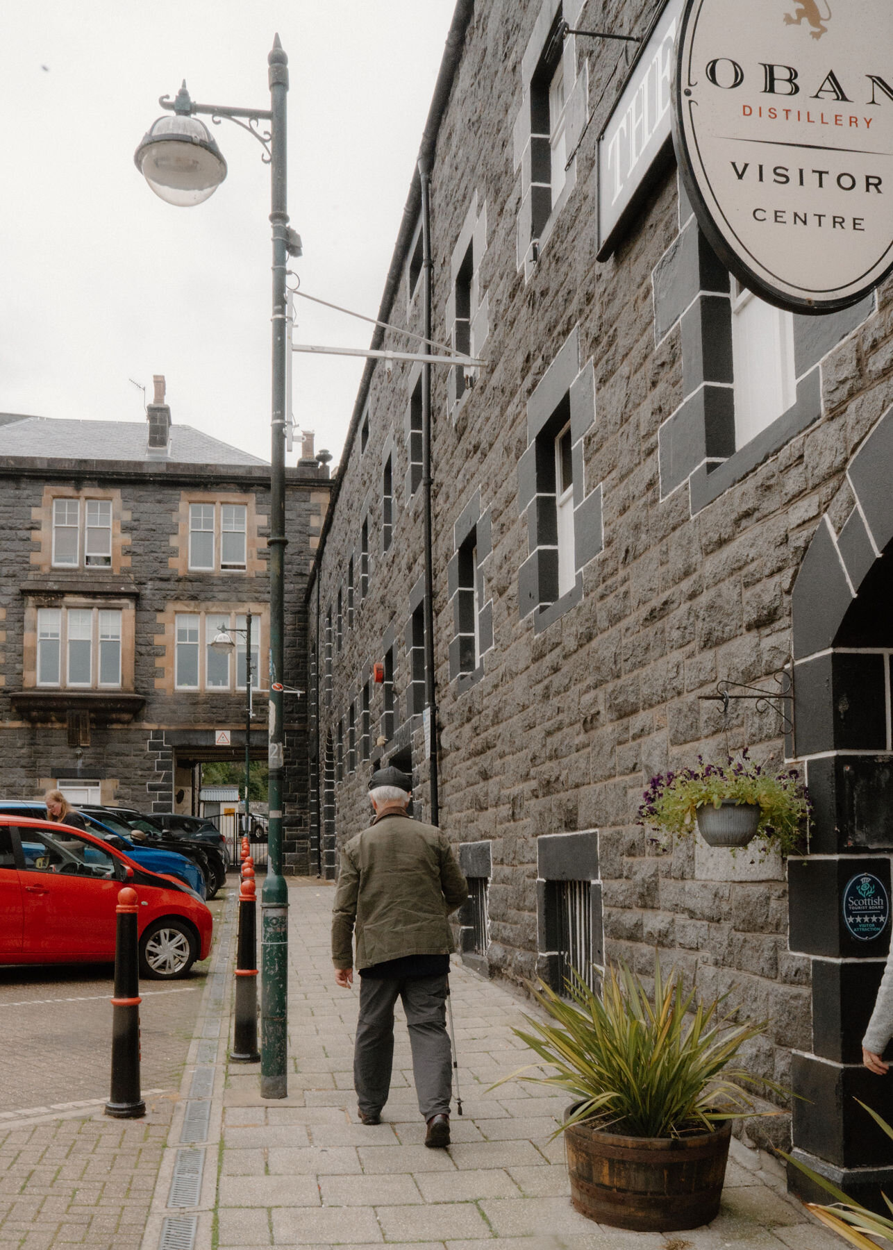 A man walks along the street past the dark stone front of Oban Distillery and its visitor centre sign.