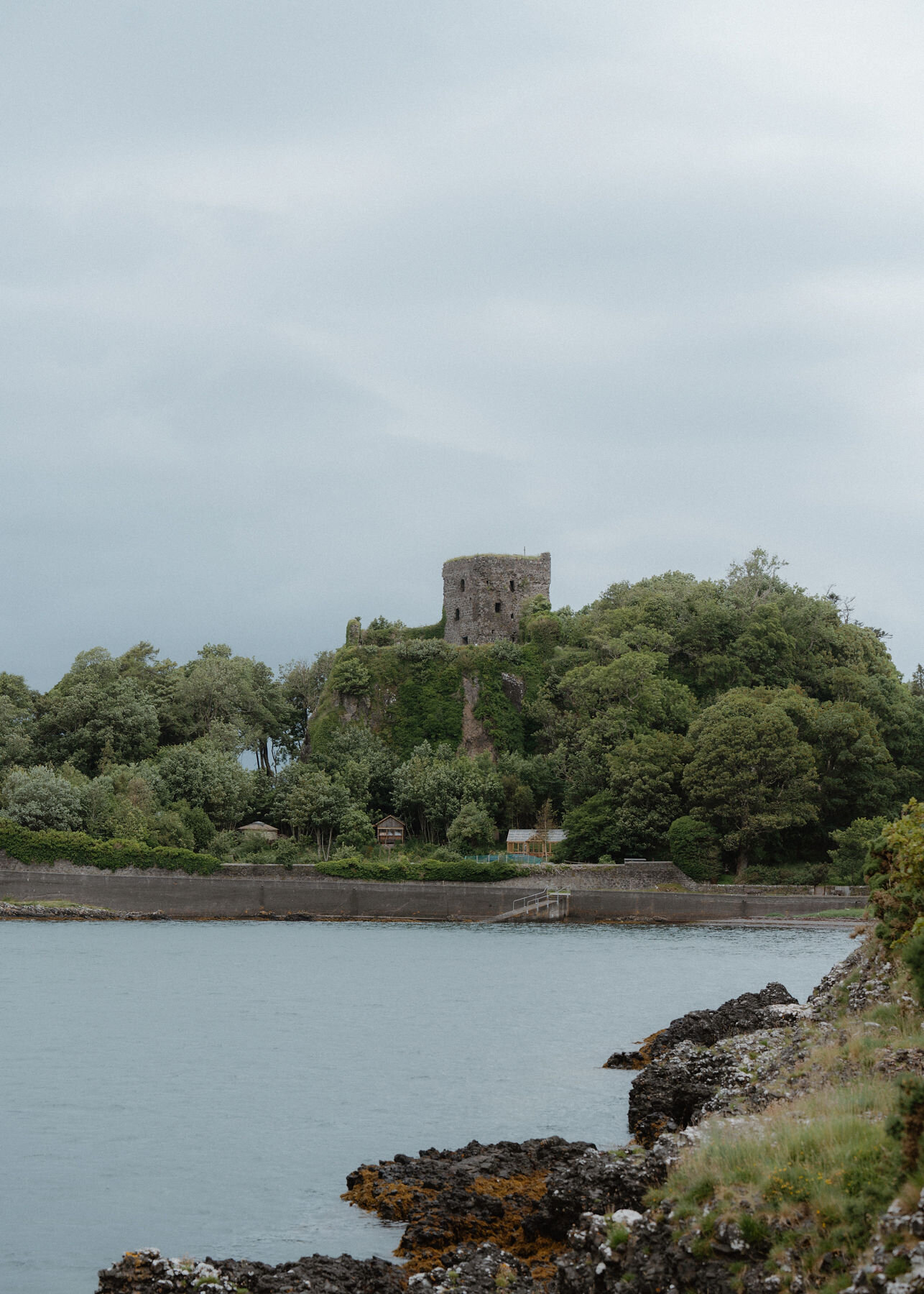 An upright frame of Dunollie's ruined keep above the trees, the calm bay and rocks in front.
