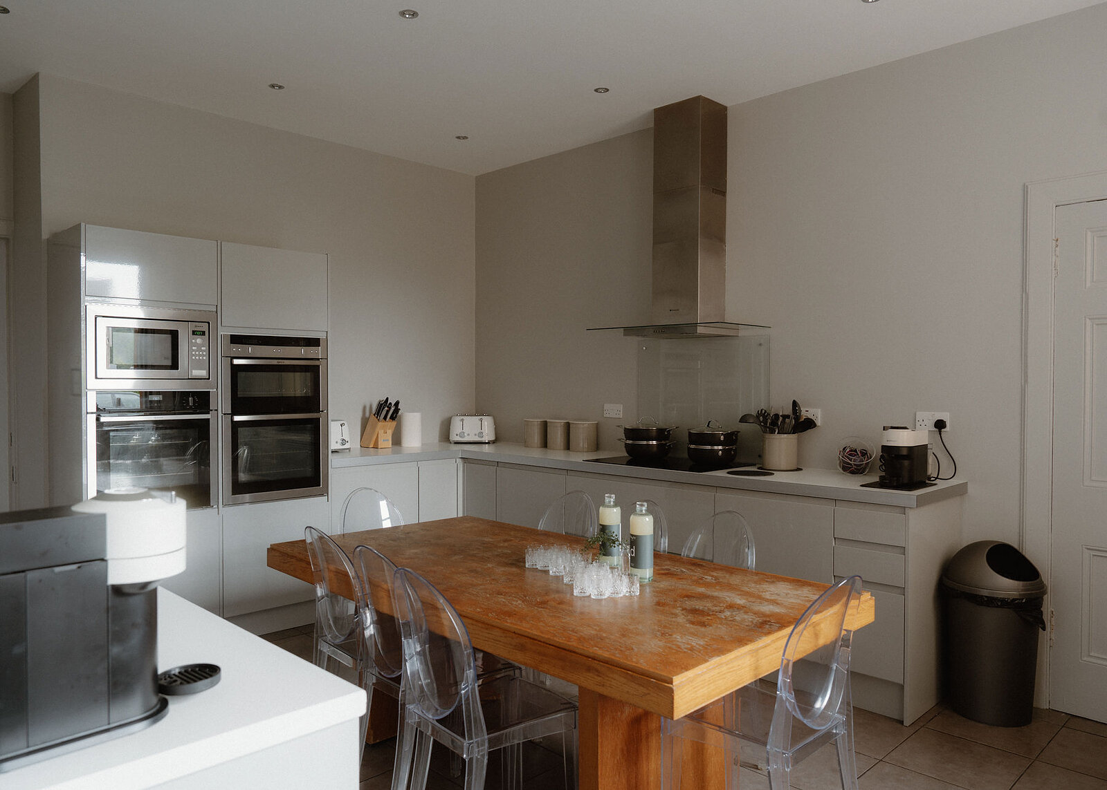 The kitchen: a long wooden table with clear ghost chairs, grey units, two ovens, an extractor hood over the hob and a window onto the garden.
