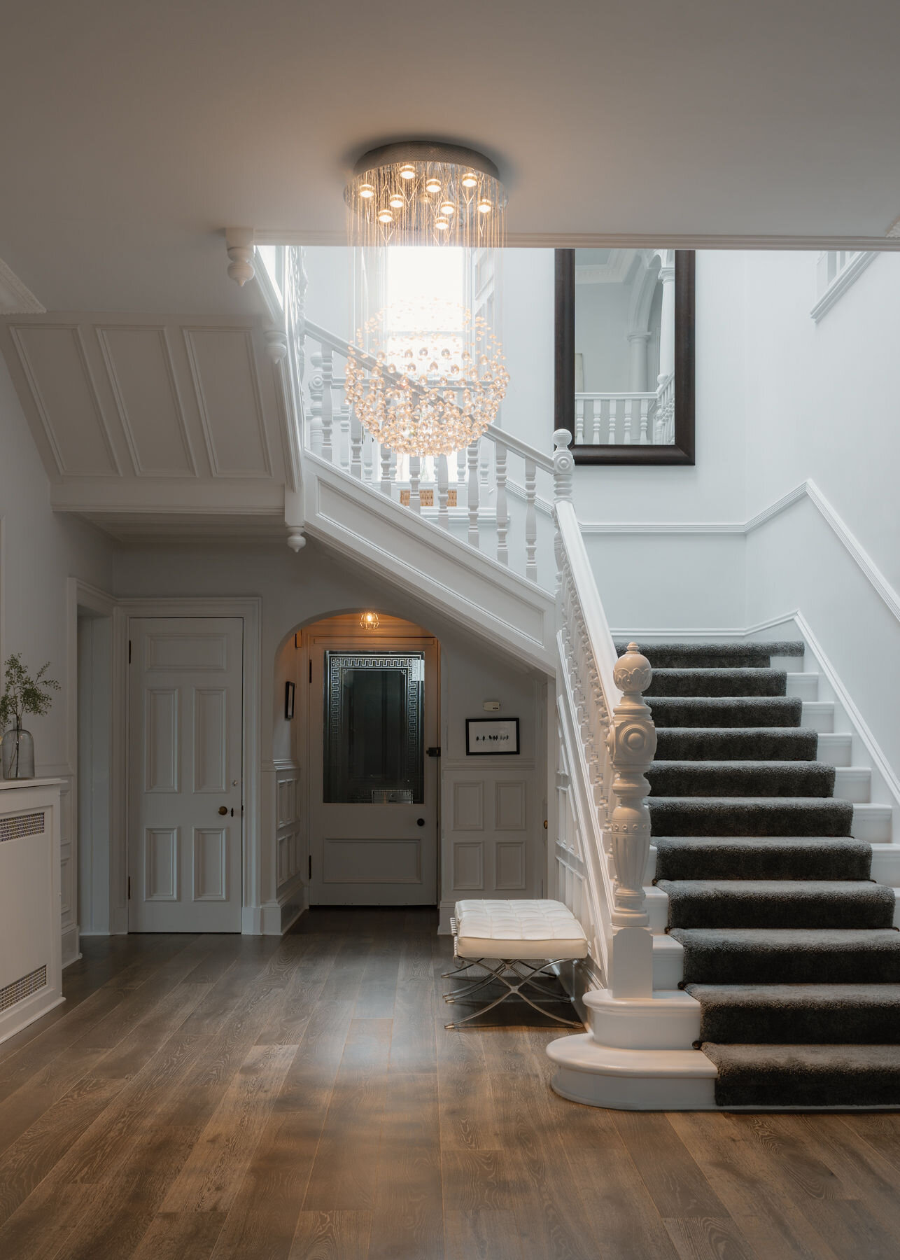 The hall: a sweeping carpeted staircase with white balusters under a cascading crystal chandelier, oak floors and an arched doorway beyond.