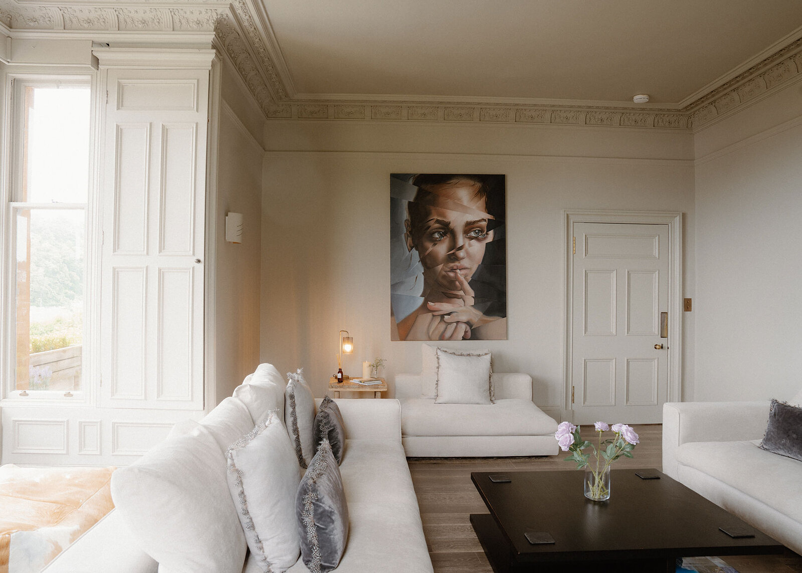 A landscape view of the drawing room: pale sofas, grey velvet cushions, roses on the table, the portrait and tall shuttered windows.
