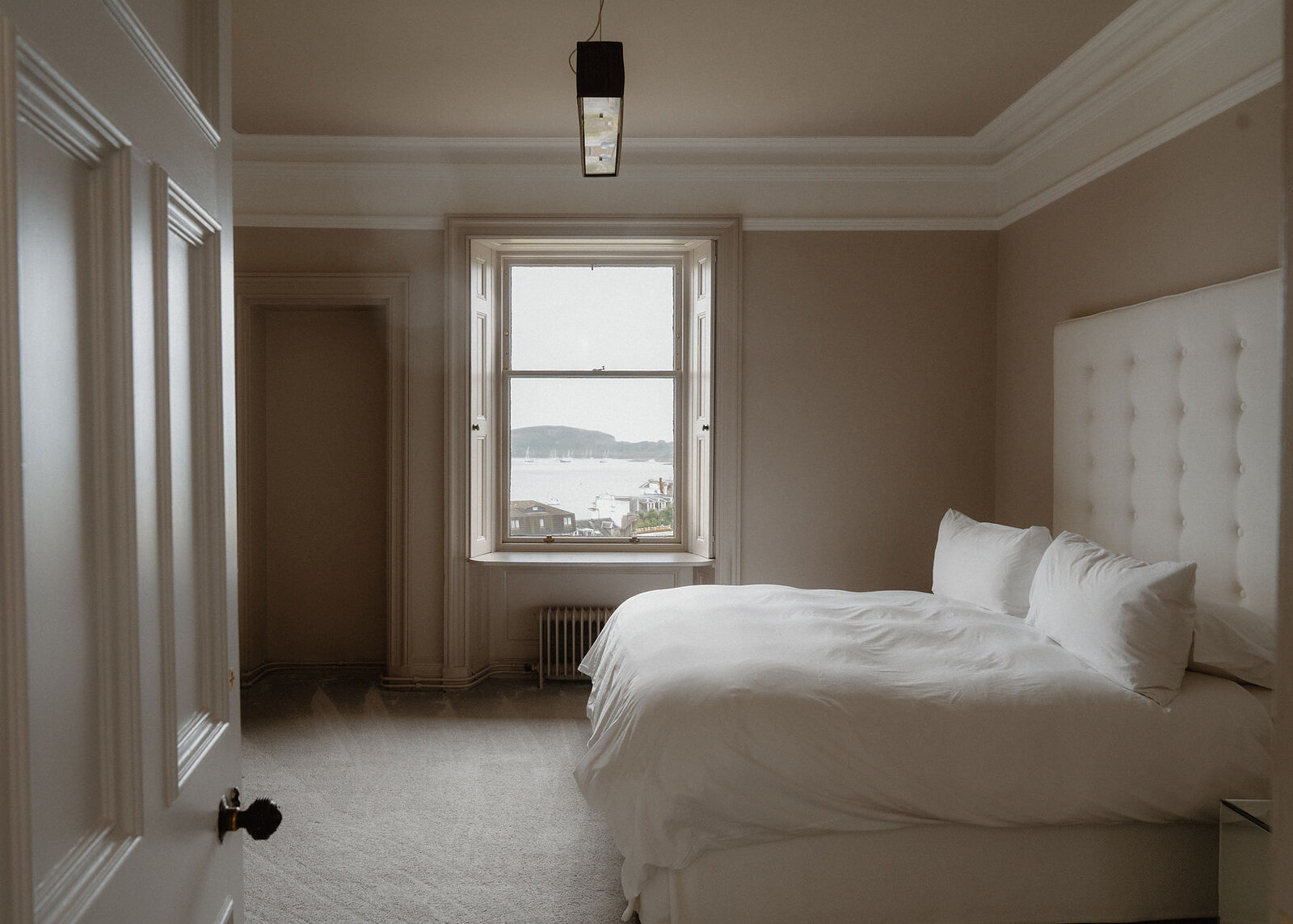 A bedroom with a tall buttoned headboard and a sash window at the end looking over the bay to Kerrera.