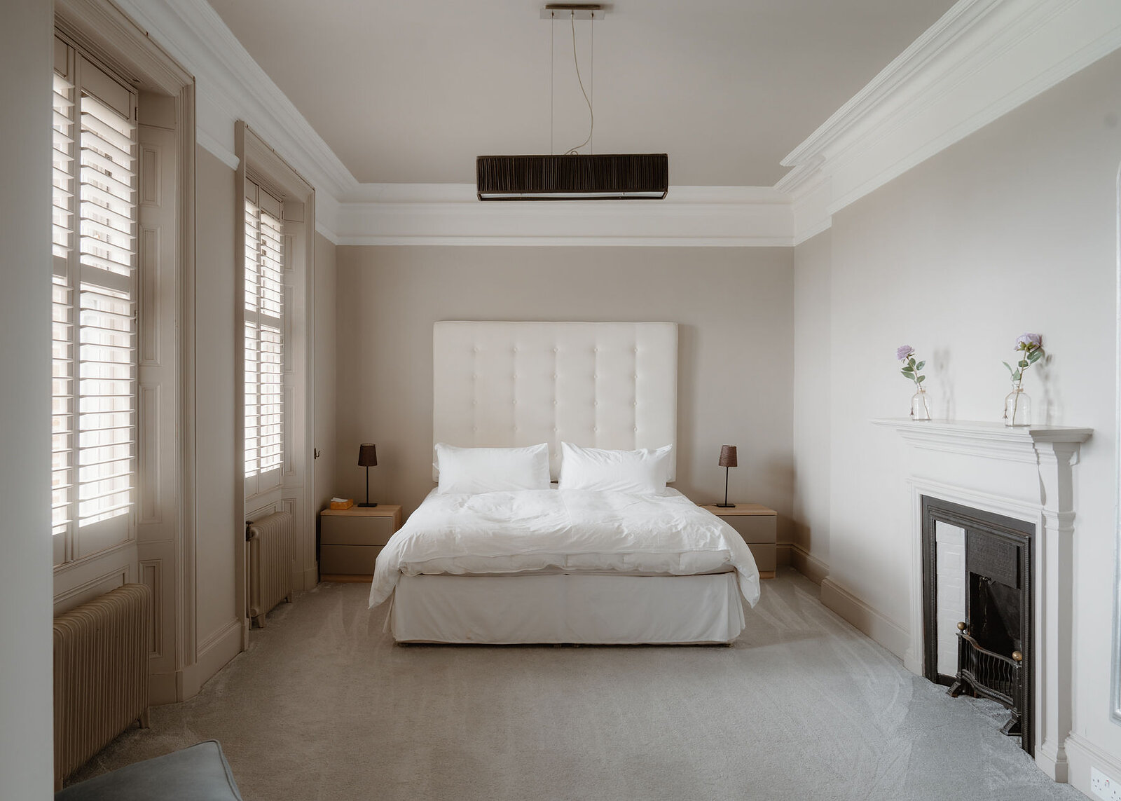 A large bedroom with shuttered windows, a white bed and a black cast-iron fireplace with flowers on the mantel.