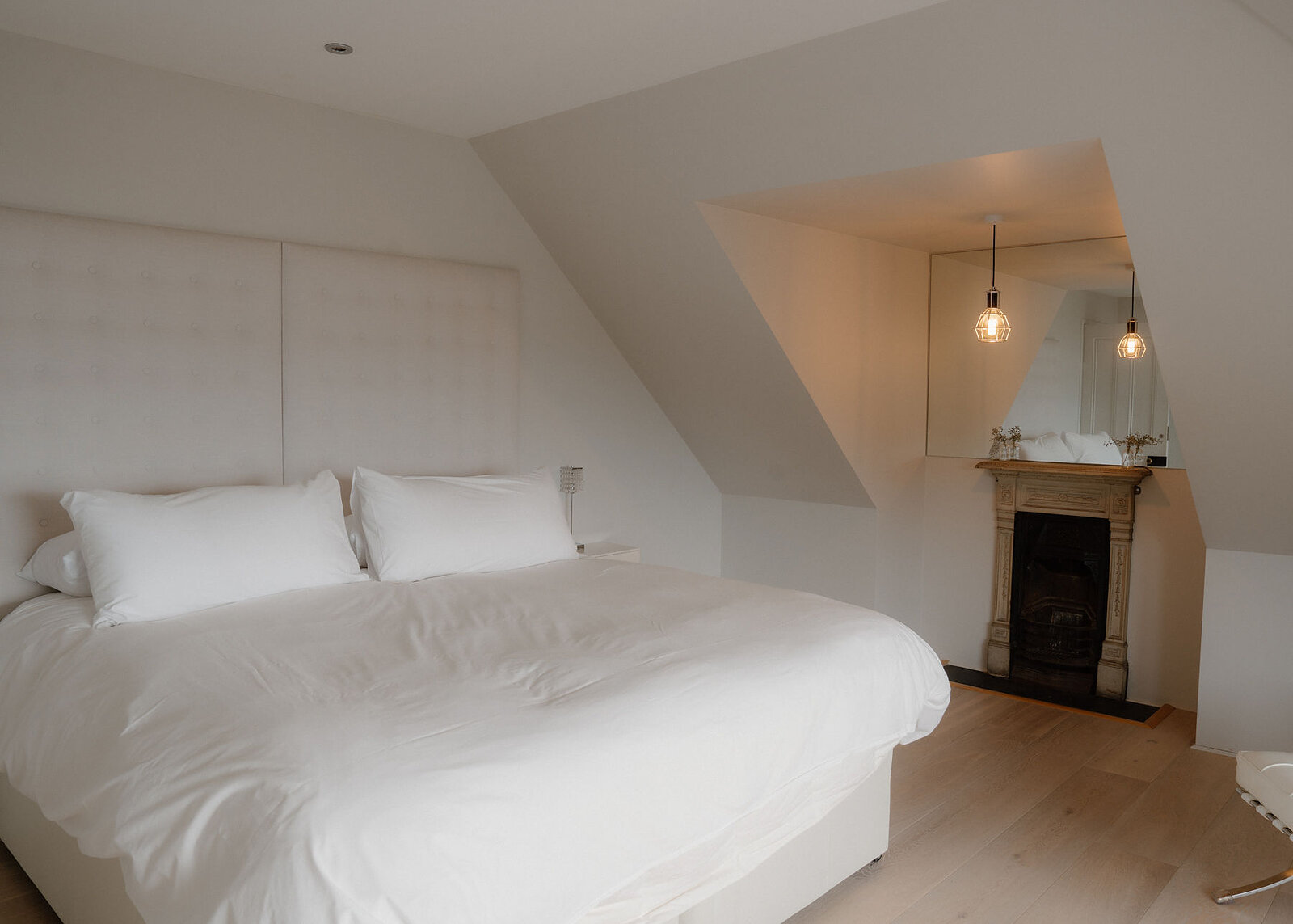 A landscape frame of the attic bedroom: a wide white bed, the old fireplace and pendant lights under the slope of the roof.