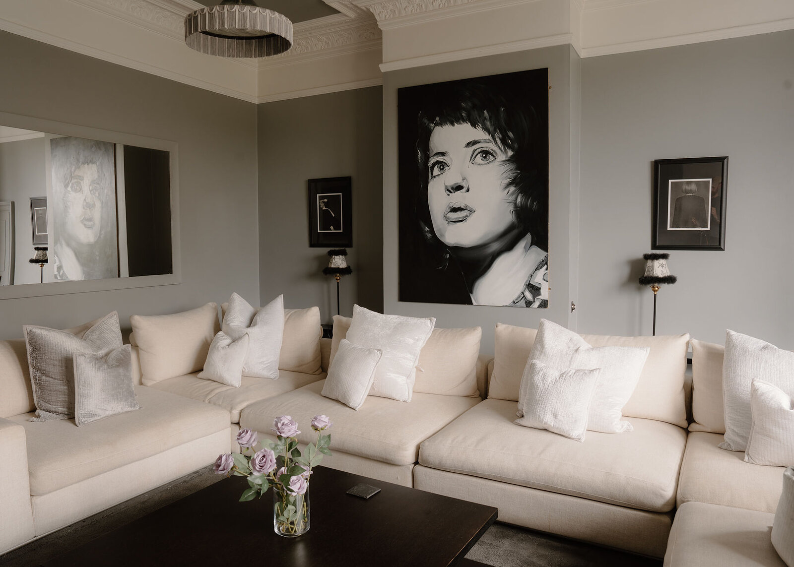 A landscape view of the cinema room: the corner sofa full of cushions, the portrait, framed pictures and roses on the table.