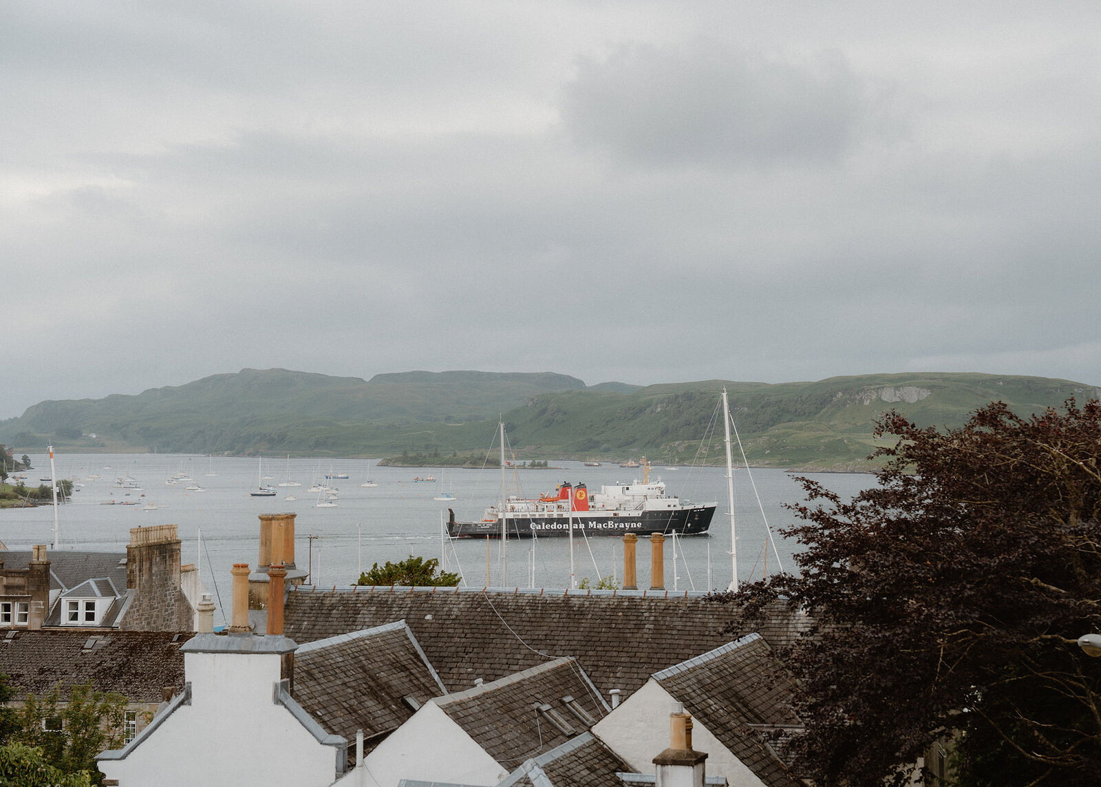 A Caledonian MacBrayne ferry crosses the bay past yachts, Kerrera beyond and Oban's slate roofs and chimney pots in front.
