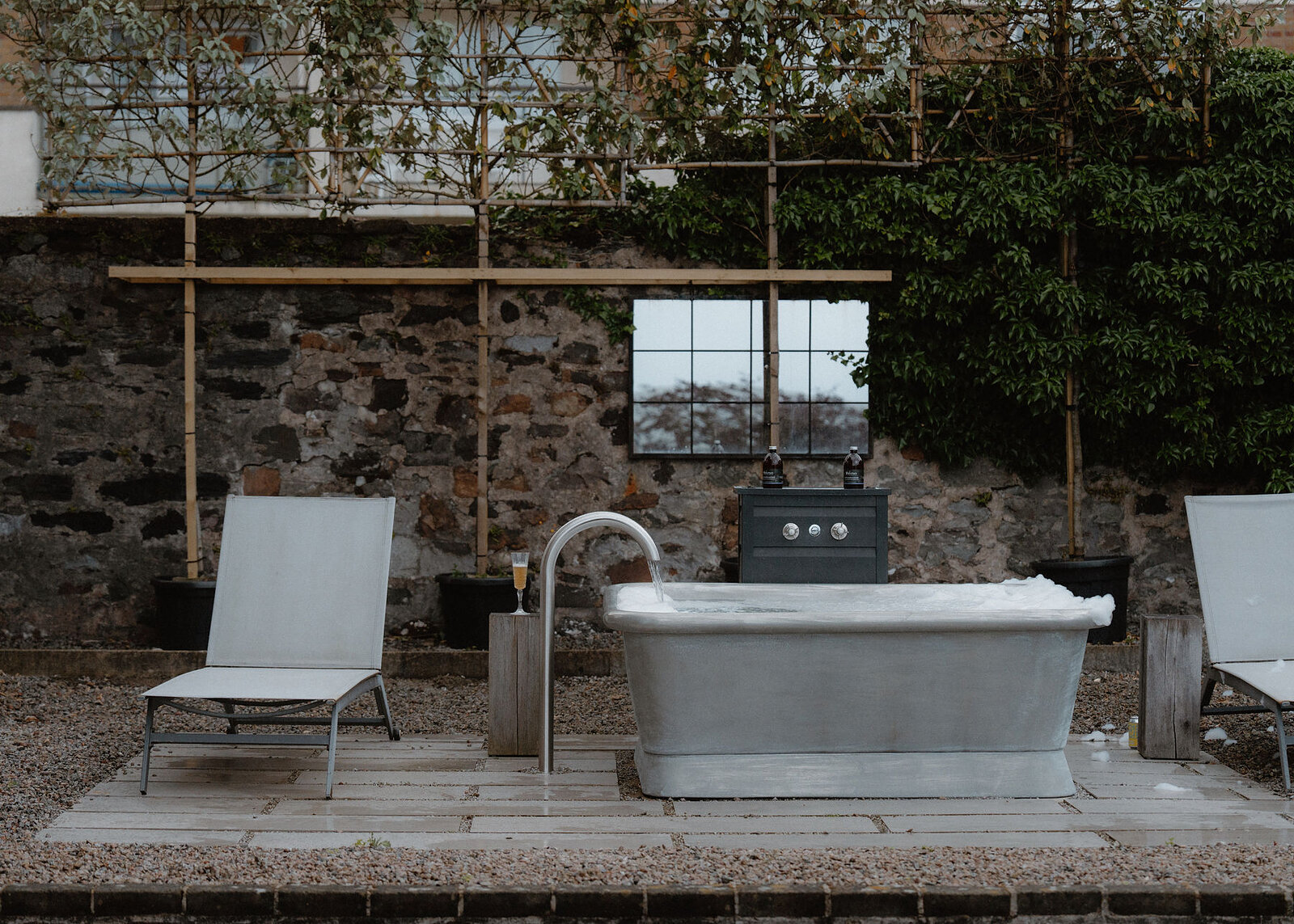 The spa bath terrace: the oversized bath between two grey loungers on stone paving, pleached trees trained along the old wall.