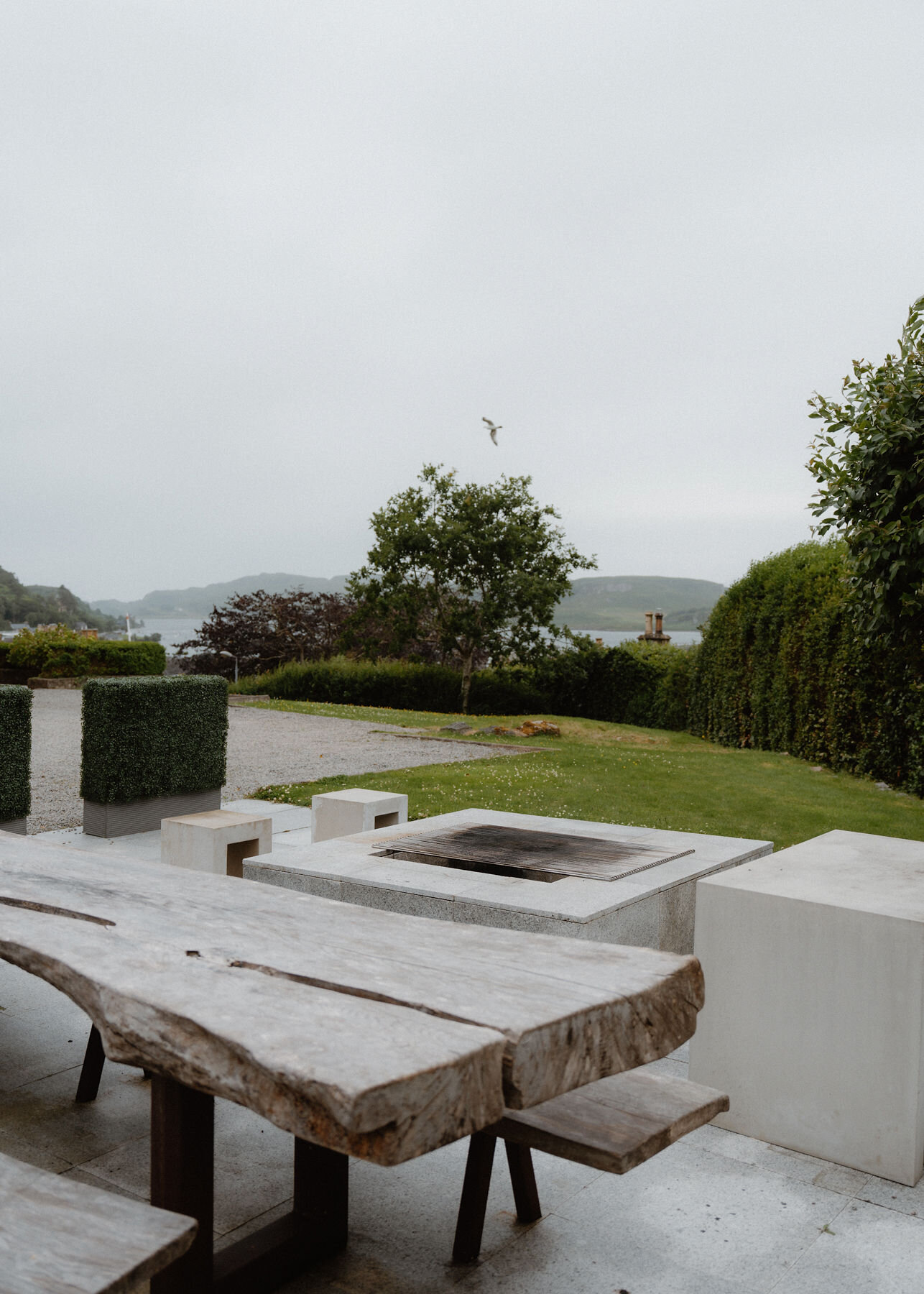 The rough-hewn outdoor table and a stone fire pit table on the terrace, the lawn and the Sound beyond.