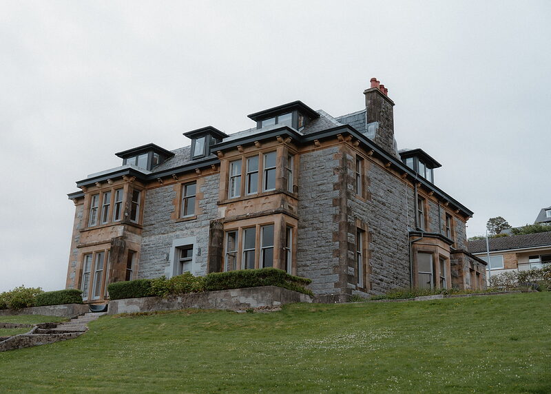 The Drury from the lawn below: a grey granite Victorian villa with sandstone bay windows, dormers in the slate roof and tall chimneys.