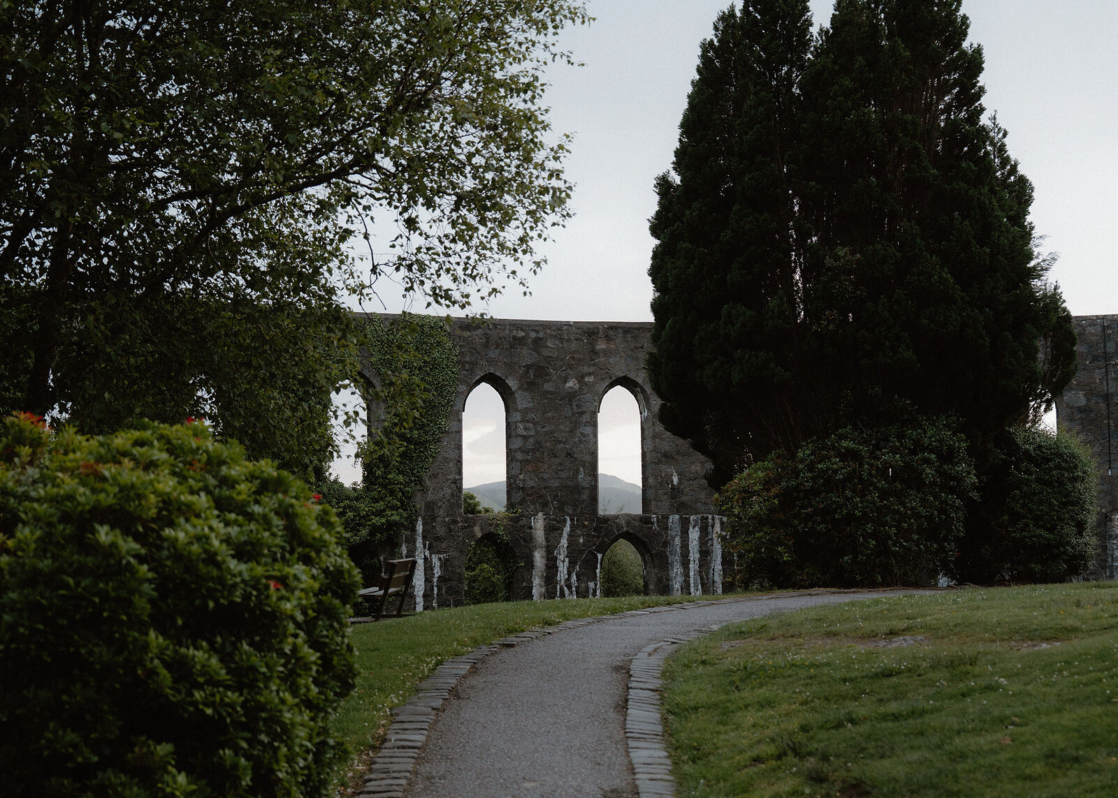 A landscape view along the path round McCaig's Tower, its tall arched openings between dark cypresses.