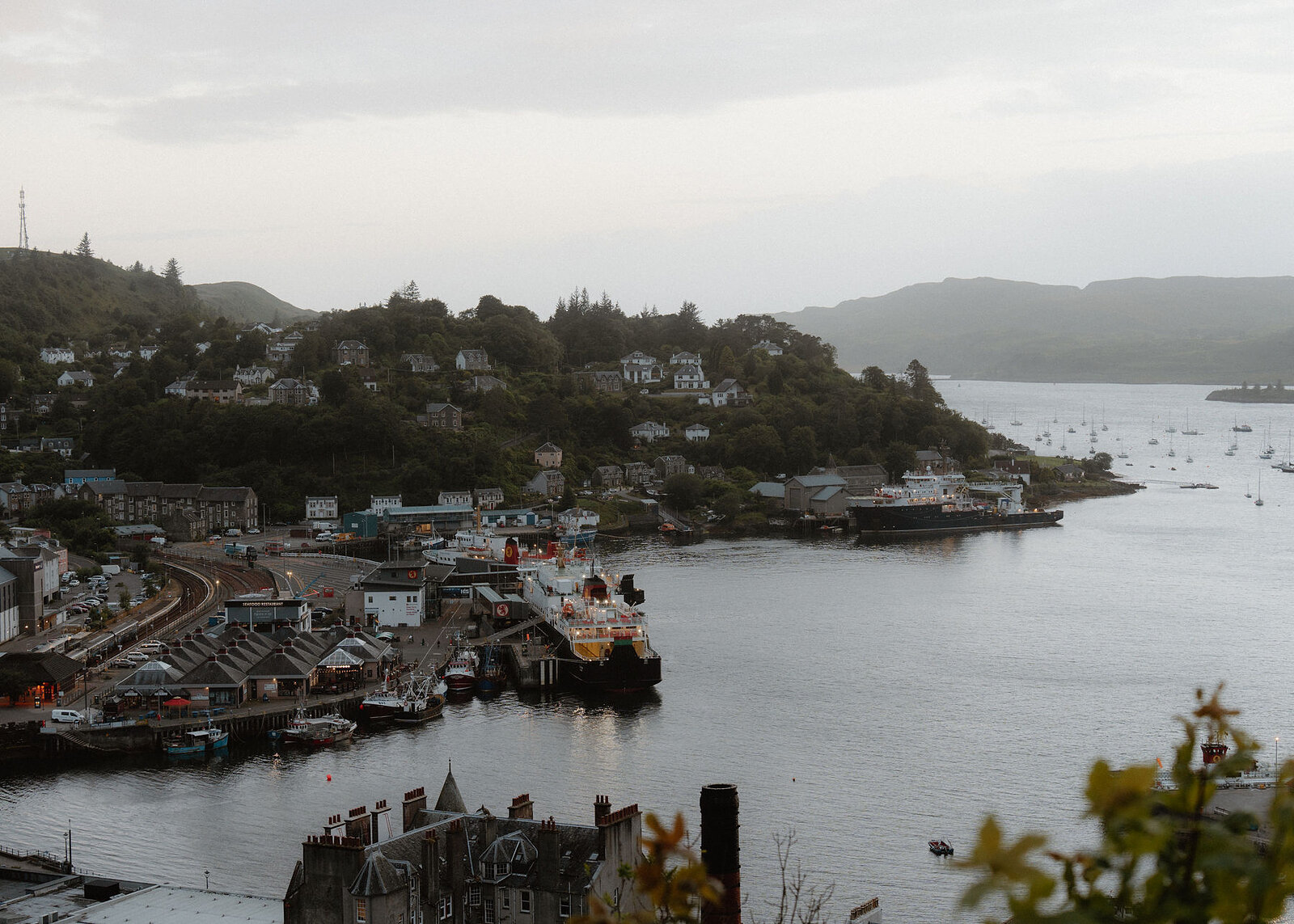 A landscape view over Oban harbour from the hill: ferries at the pier, the station and the houses rising to the north.