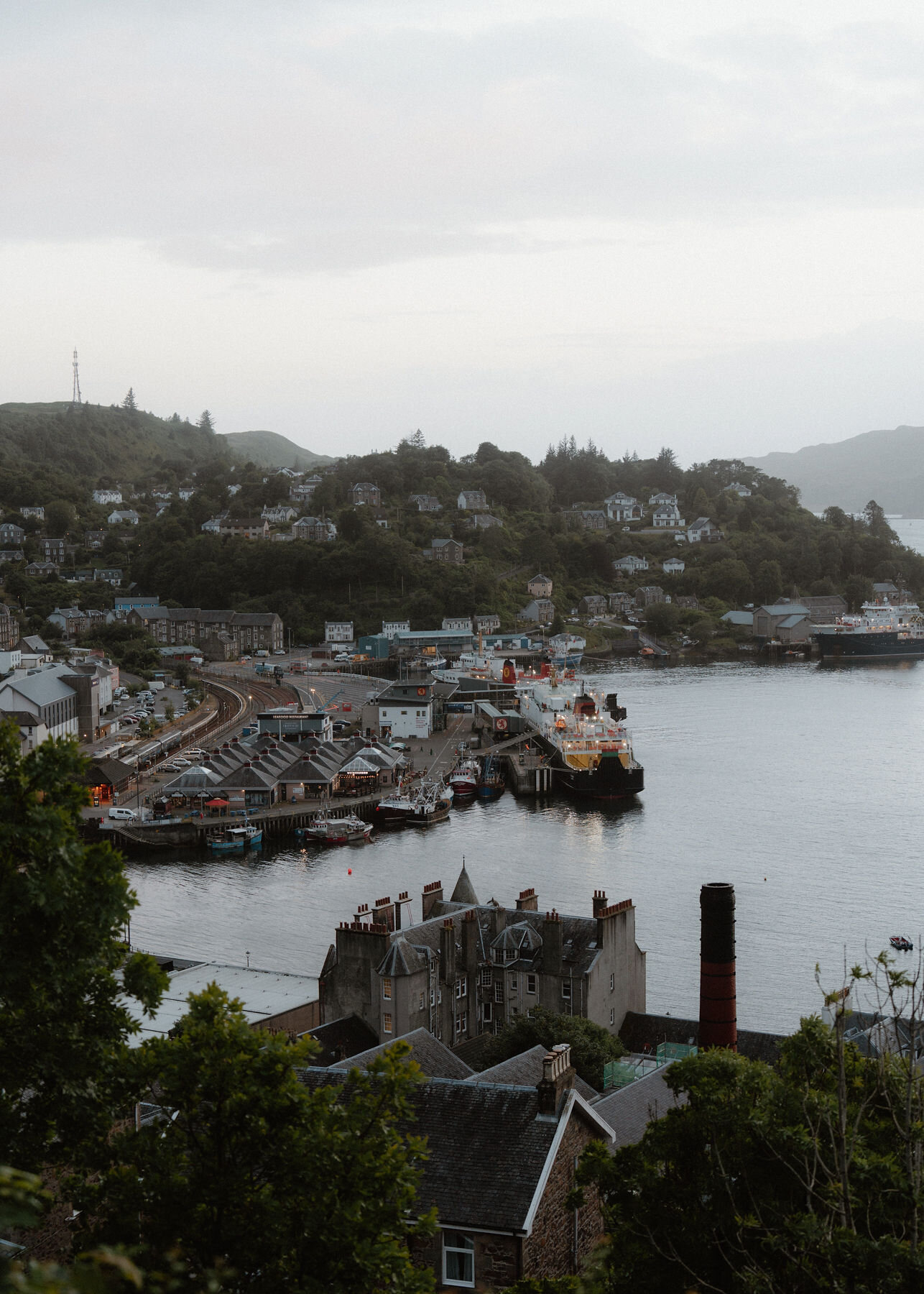 An upright frame of the harbour and ferry terminal below the hill, the distillery chimney in the foreground.