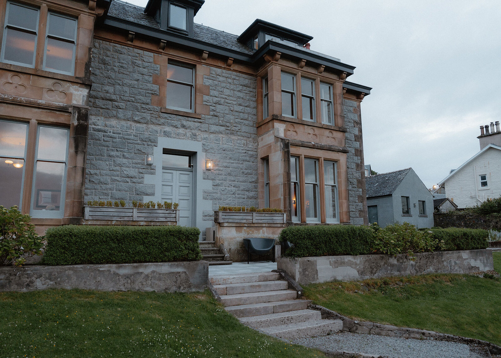 A landscape view of the house's front at dusk: the door and its lamps, the bays, the steps and the clipped hedges.