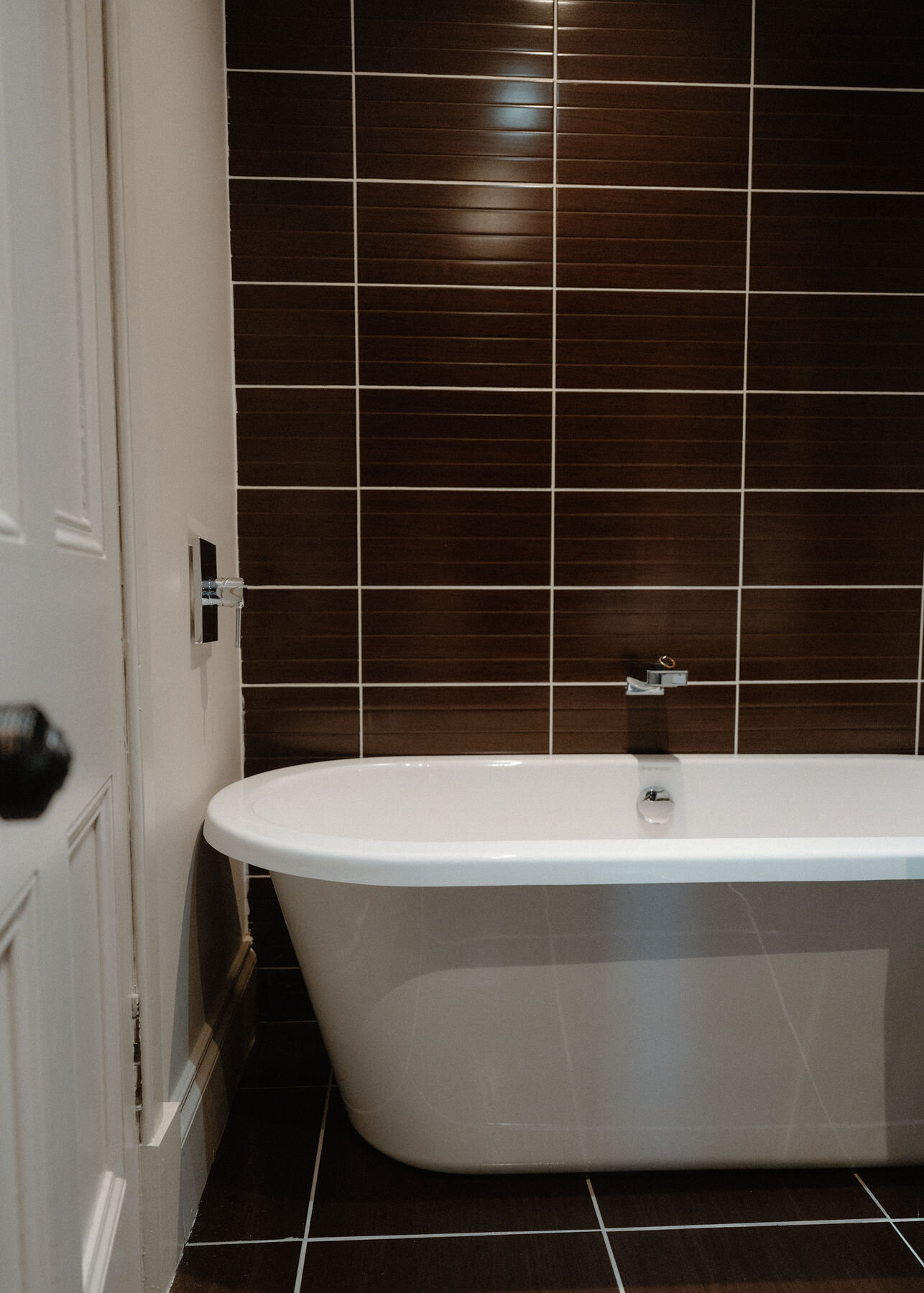A freestanding white bath in a bathroom tiled in glossy dark brown.