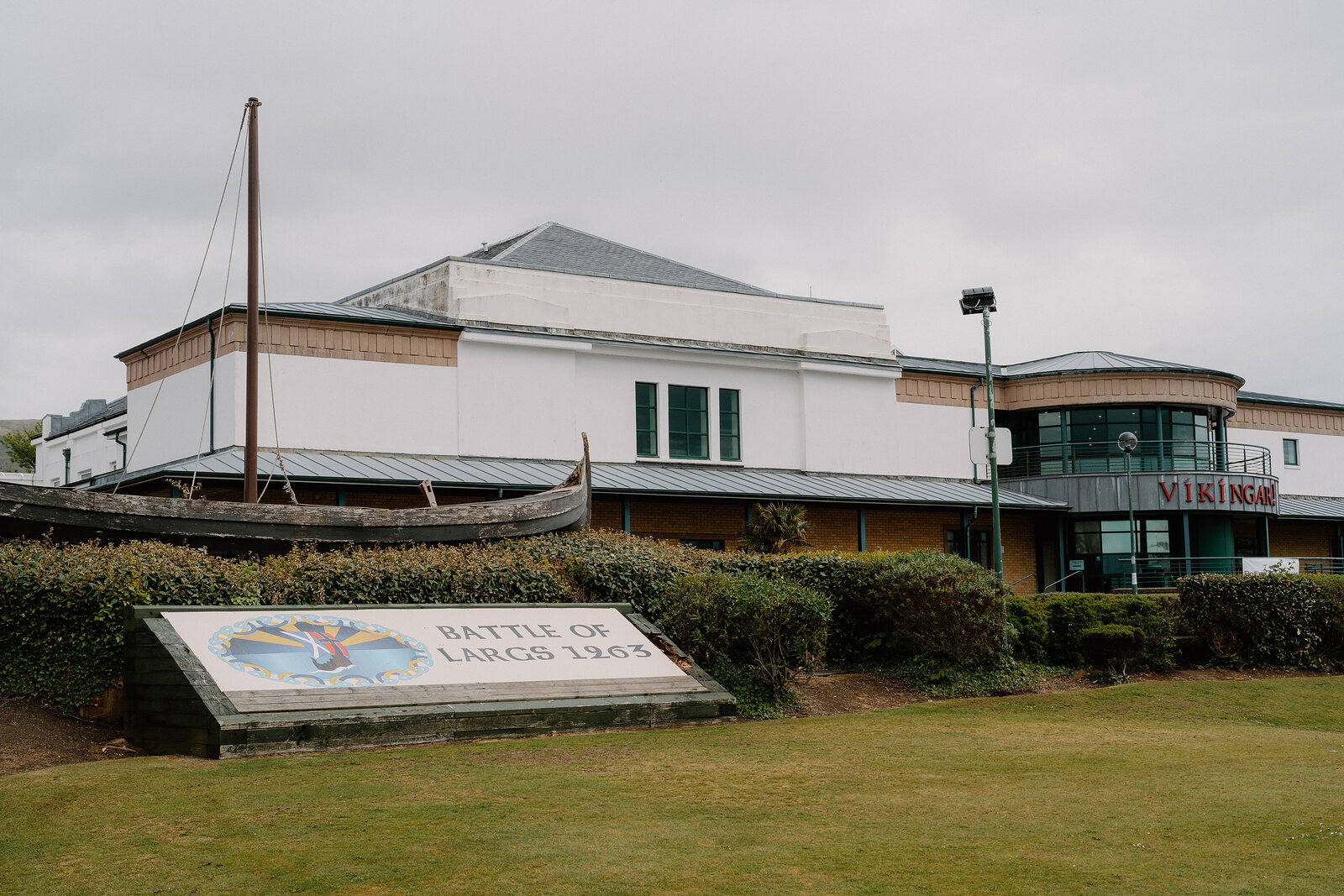 A replica Viking longship stands beside a building with a rounded gable, a stone monument inscribed "Battle of Largs 1263" on the lawn in front.