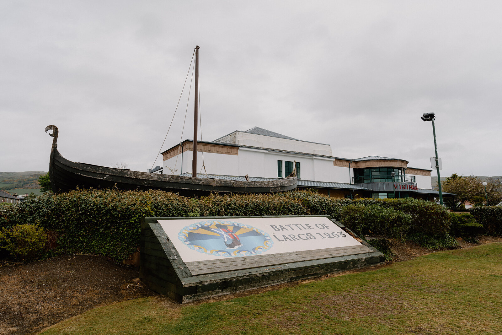 A replica Viking longship stands beside a curved building, a stone monument inscribed "Battle of Largs 1263" on the lawn in front.
