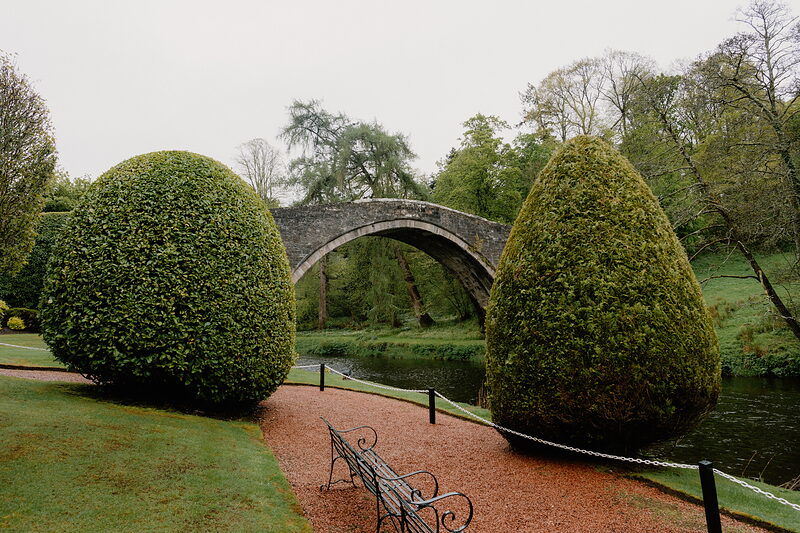 An old stone humpback bridge is framed between two dome-clipped yews, a bench standing on the gravel path beside the river.