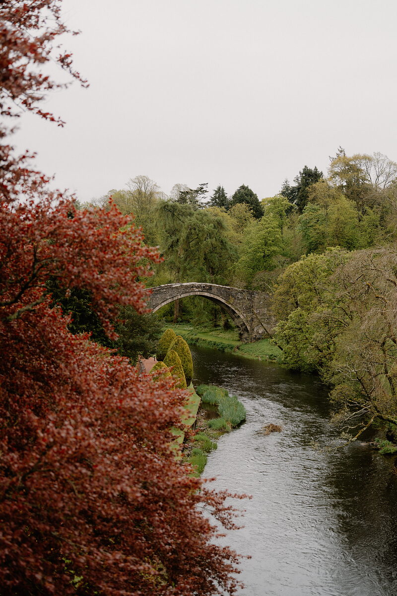 An old stone humpback bridge is glimpsed through autumn beech trees, a river winding below between wooded banks.