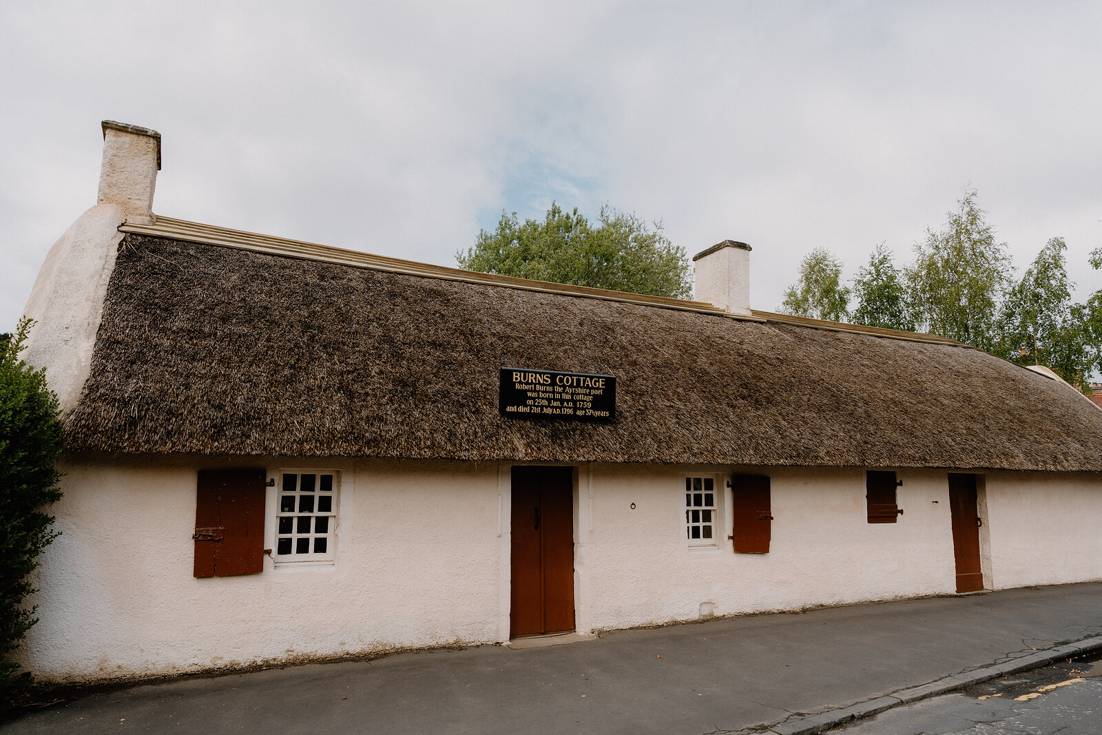 A long whitewashed thatched cottage signed Burns Cottage stands along a street, red shutters and doors breaking its plain front.